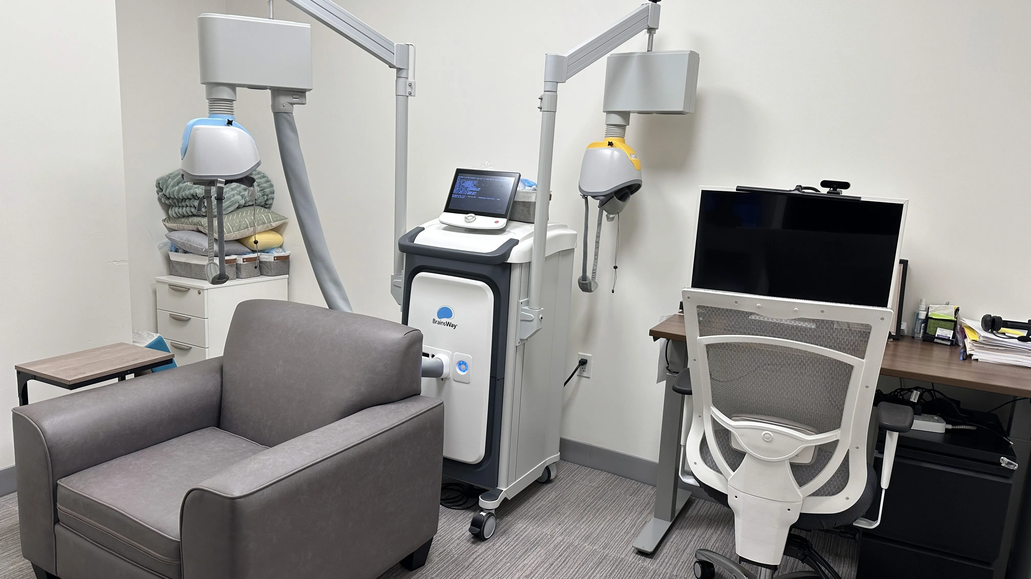 Two Deep TMS treatment devices with coils beside a desk with a computer monitor and office chair