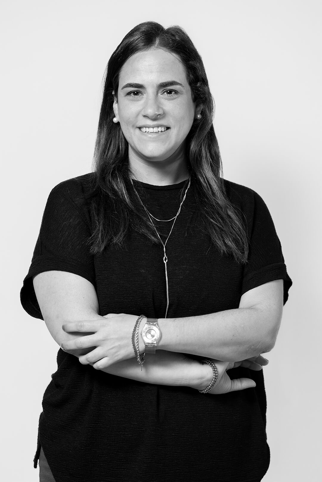 Black and white image of a person smiling warmly with arms crossed, wearing a black top, layered necklaces, bracelets, and a watch, conveying confidence.