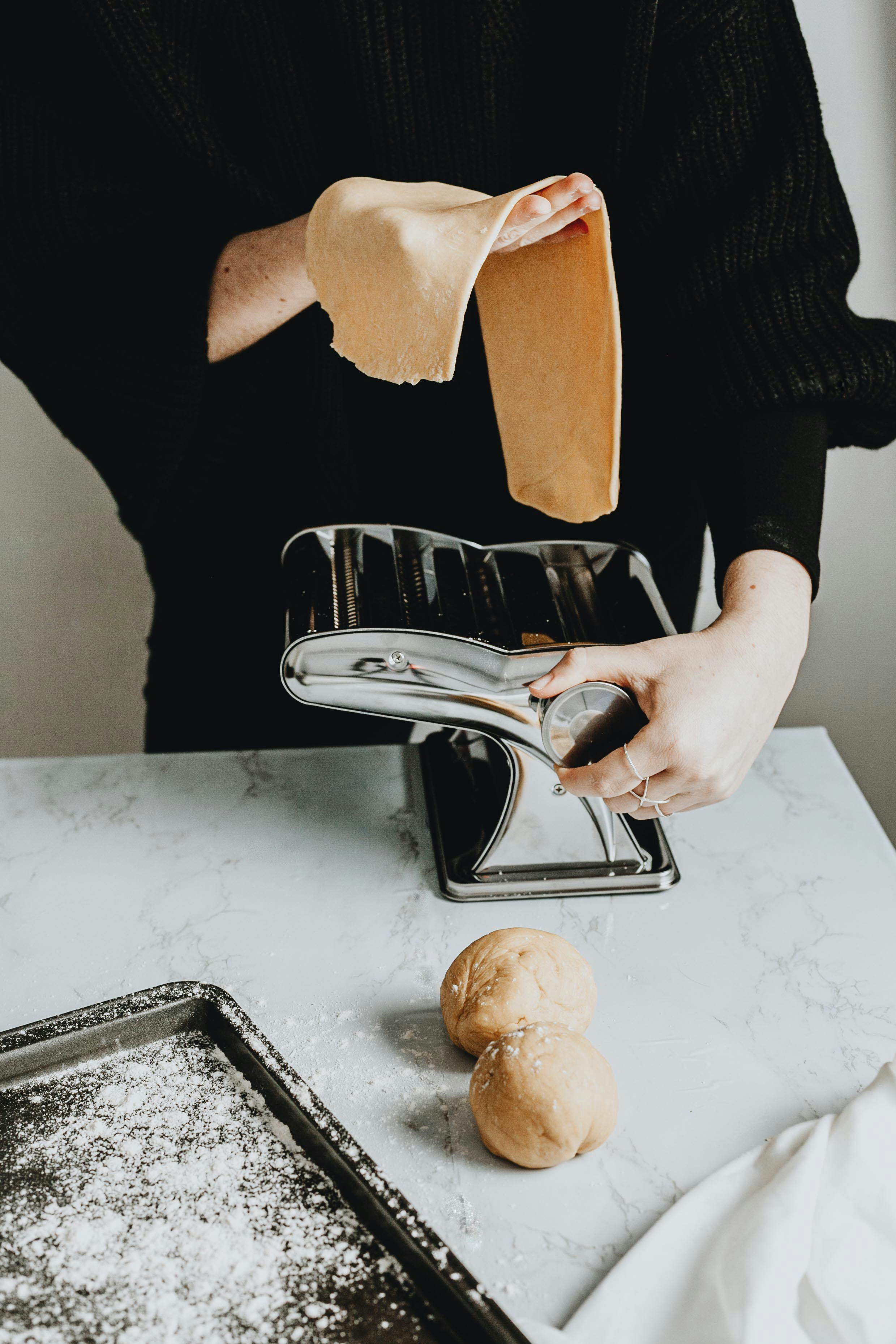 A person wearing a black sweater uses a stainless steel pasta maker on a marble countertop, with rolled-out dough being fed into the machine and two dough balls nearby, set against a softly lit kitchen background.