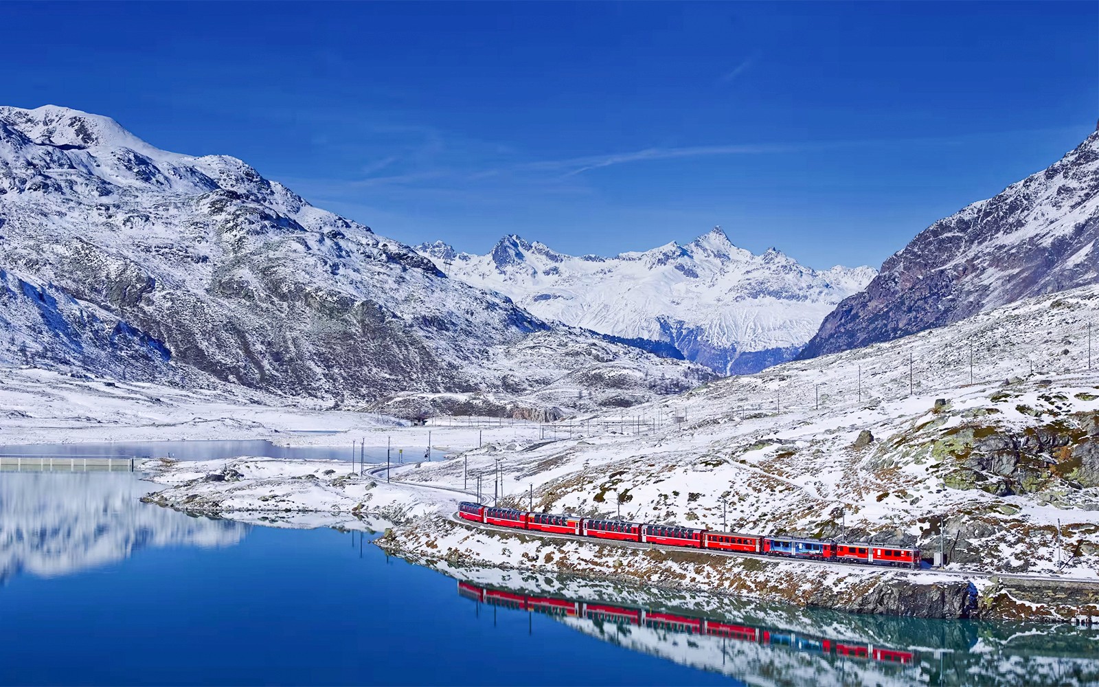 Bernina Ekspresi treninin karlı İsviçre Alplerinde göl üzerindeki dağ yansımasıyla yolculuğu.