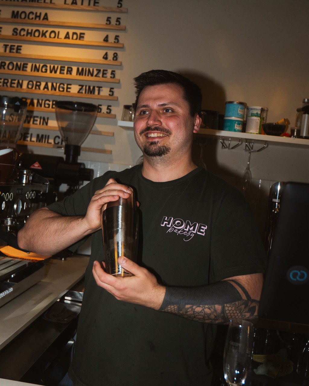 A bartender with a tattooed arm skillfully shakes a cocktail shaker behind a counter, surrounded by coffee and drink equipment, under a menu board with various beverage options.