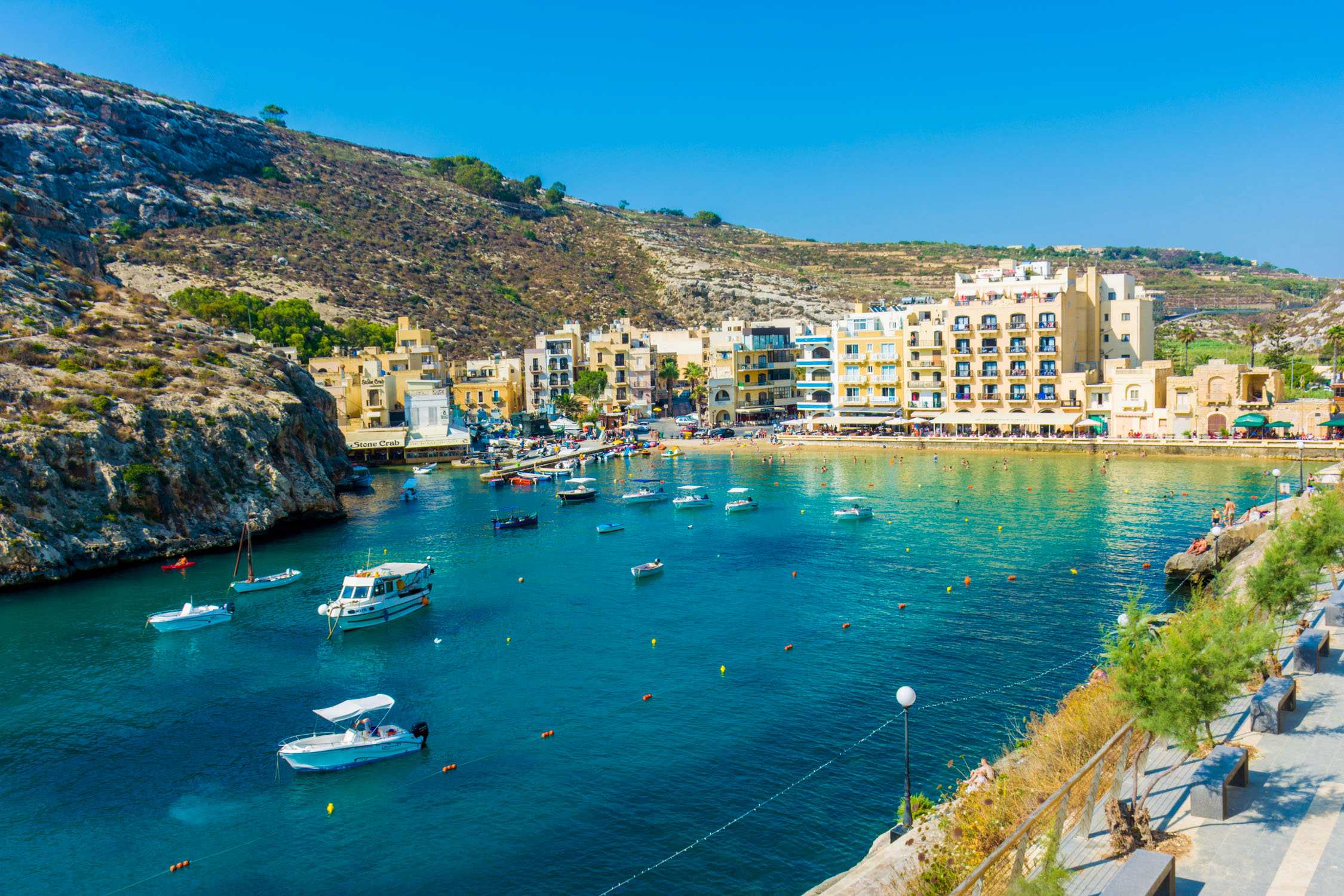 Bright sunlight illuminates a turquoise bay nestled between rocky hills and buildings lining the waterfront promenade.