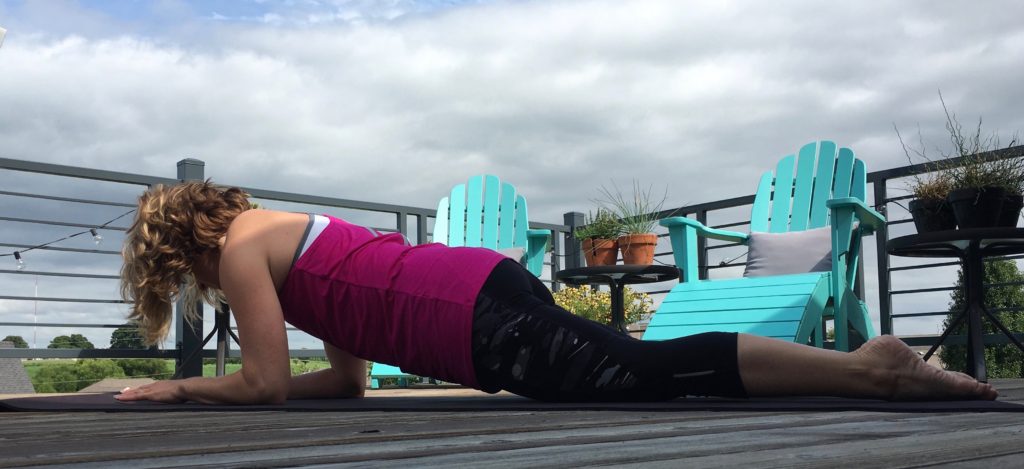 Heather Hausman practices yoga on a wooden deck under a cloudy sky, wearing a pink tank top and black leggings, with turquoise Adirondack chairs and potted plants in the background.