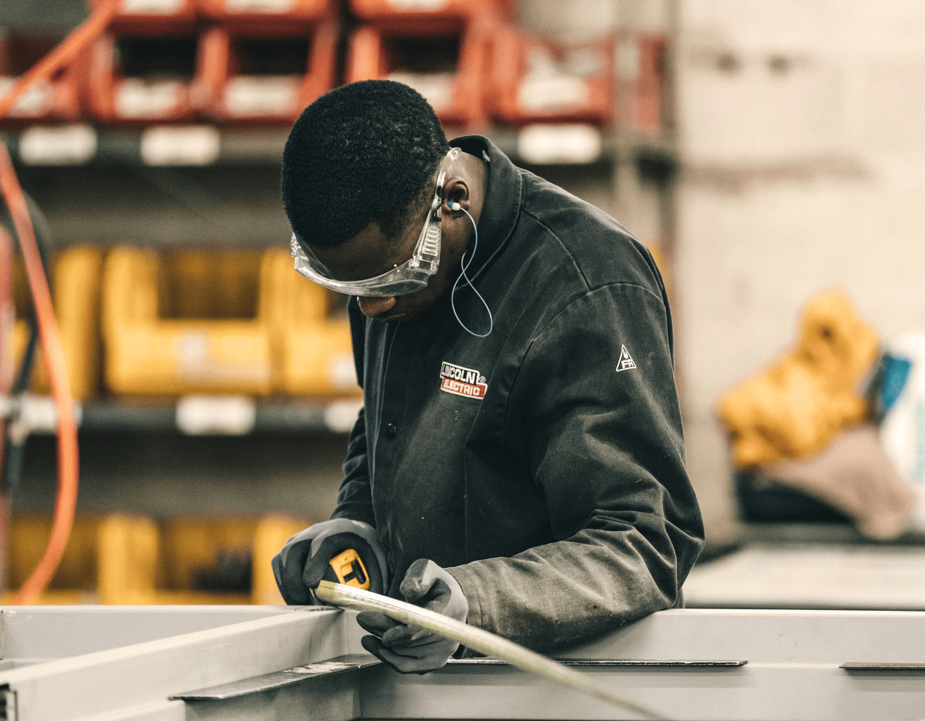 A person in work attire focuses on assembling metal components in a workshop filled with tools and materials.