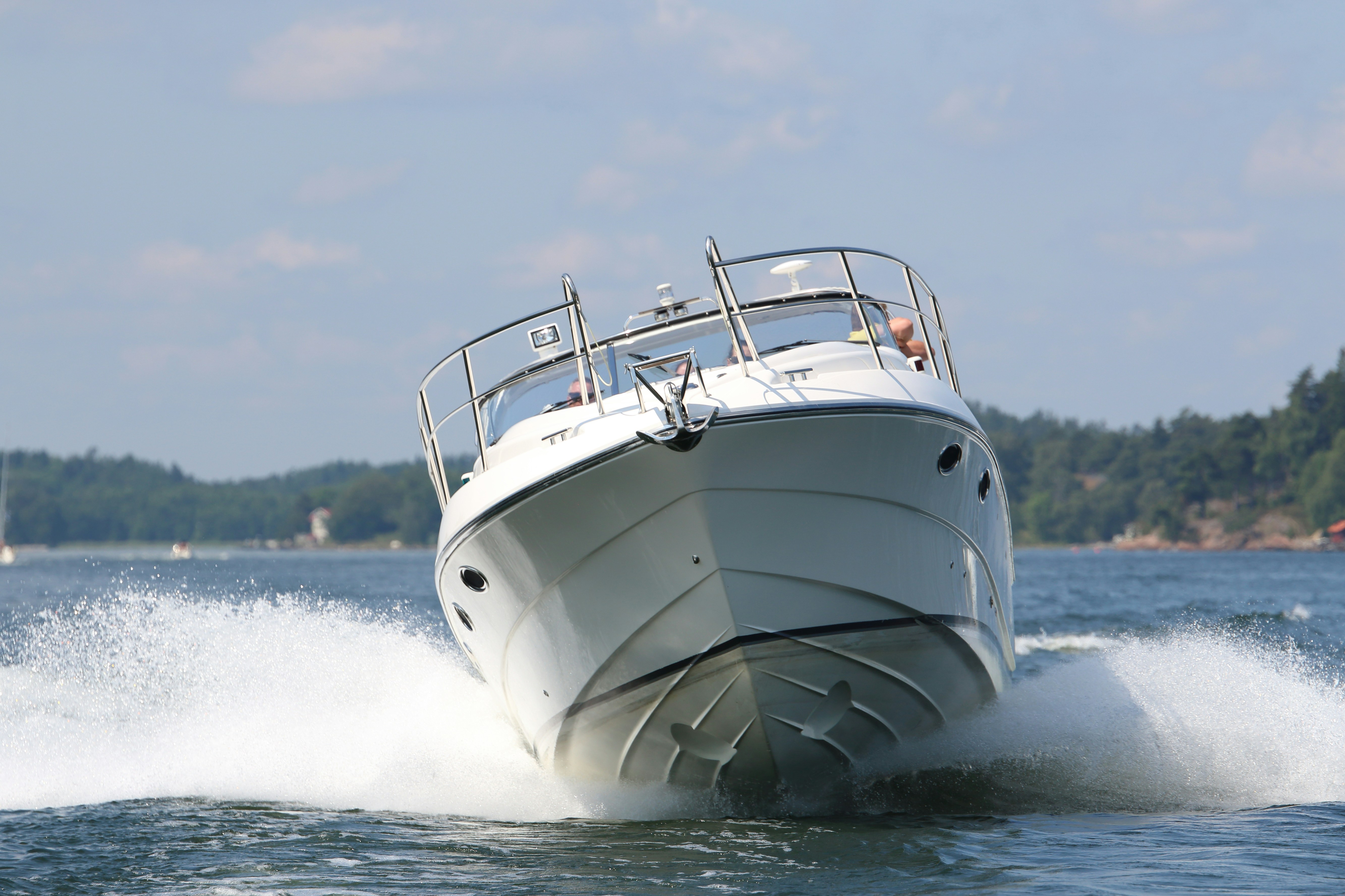 A sleek white motorboat cuts swiftly through the blue waters, creating a frothy white wake under a clear sky, with lush green forested islands in the background.