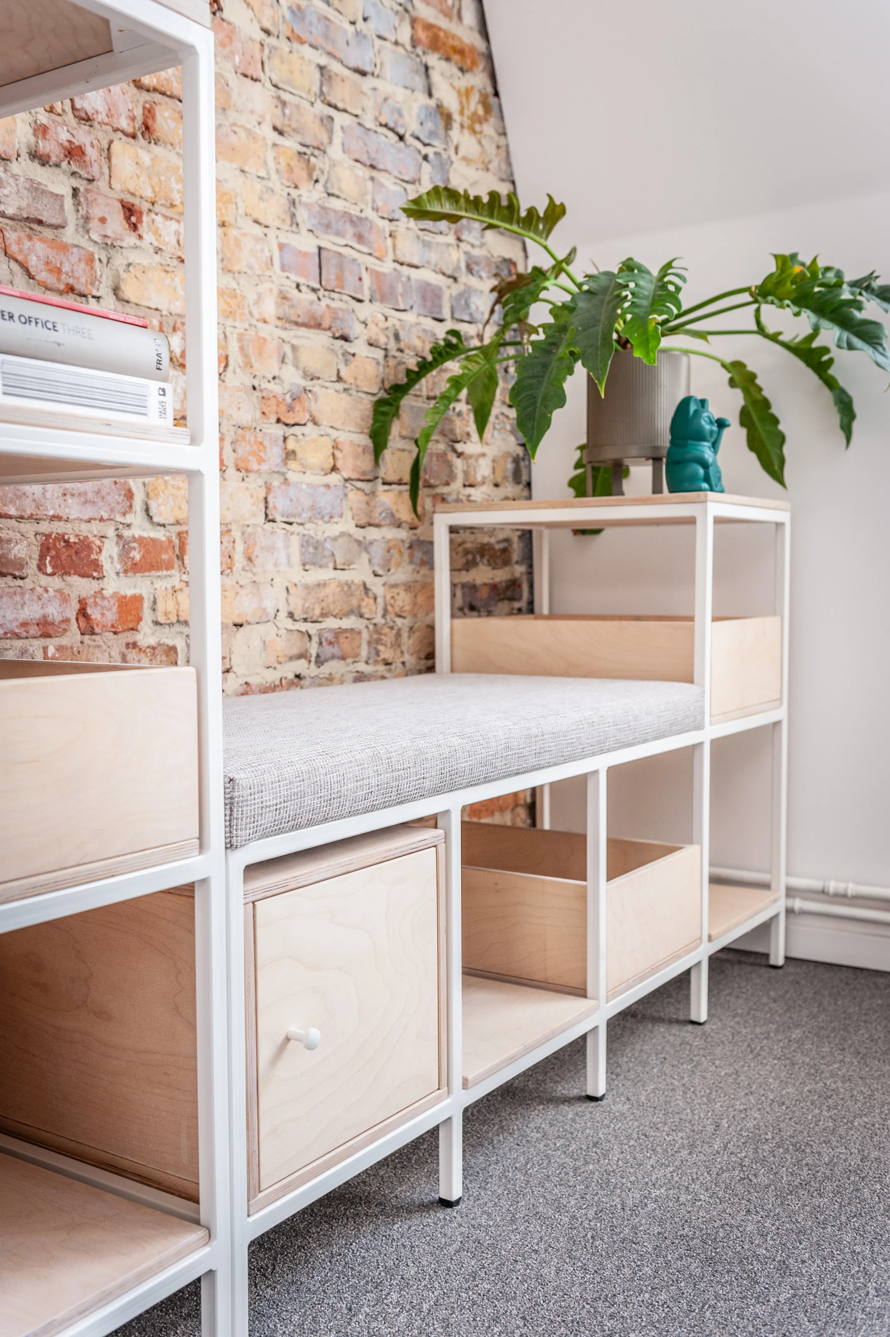White steel shelving unit at EHEALTH-TEC with birch plywood boxes and a grey upholstered bench cushion in front of an exposed brick wall, topped by a large philodendron plant.