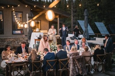 Wedding guests gather around an outdoor dinner table beneath string lights beside the cabin.