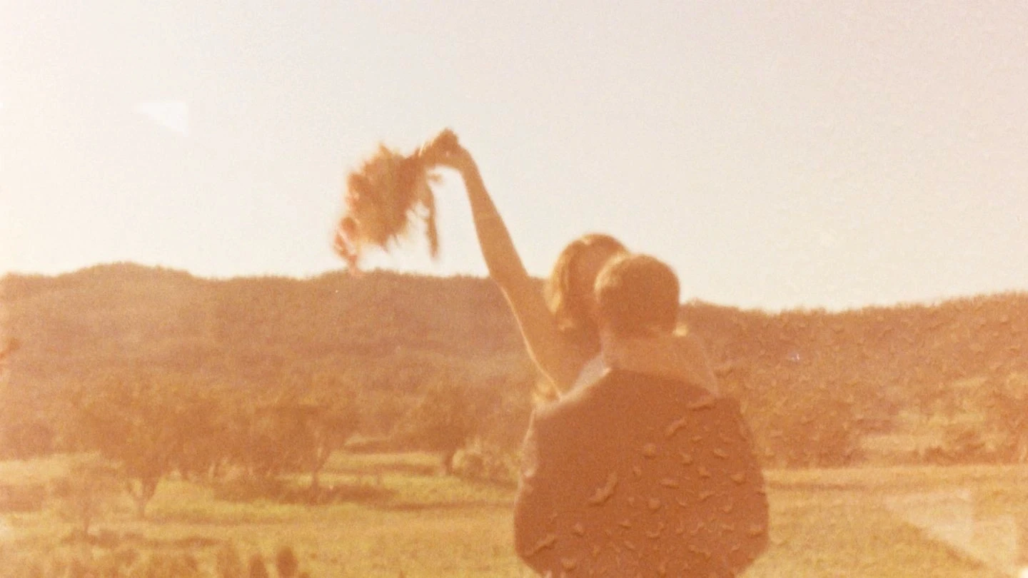 A couple embraces joyfully, with the woman raising a bouquet against a warm, sunlit rural landscape backdrop, capturing a moment of celebration and romance.
