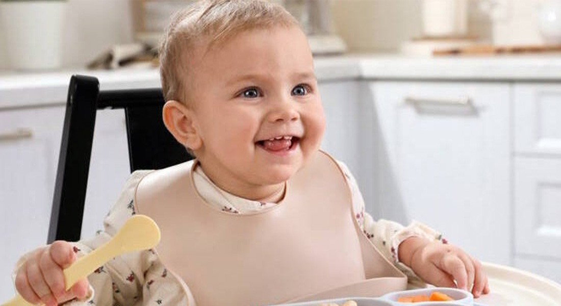 Kisgyermek etetőszékben ülve kivi szeletet harap, kíváncsian nézve oldalra.