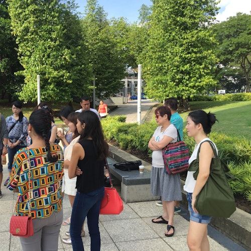 People standing outdoors in a park, some chatting and others looking around. Greenery and a paved path are visible.