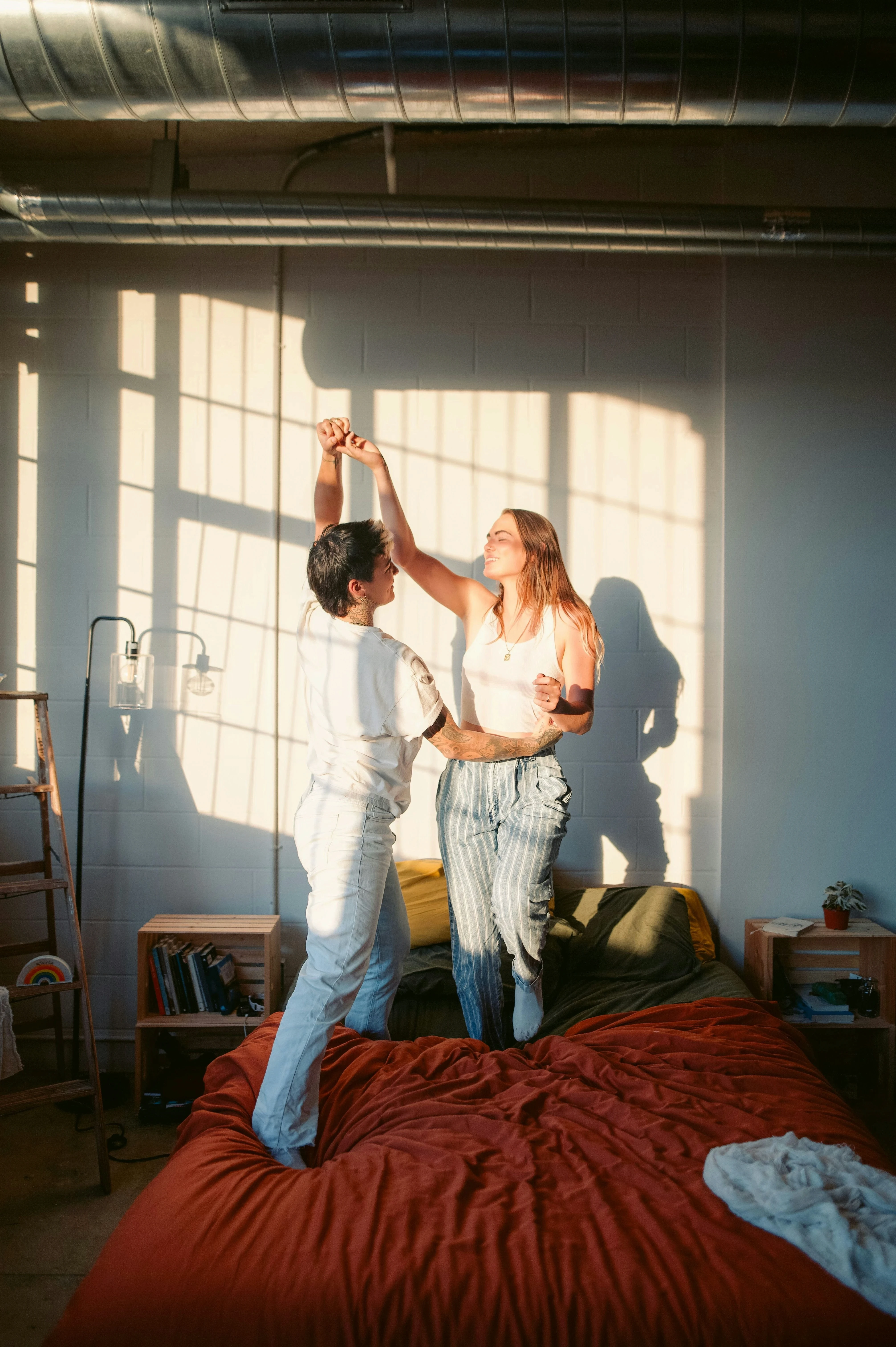 Two queer women dancing together on a bed in a sunlit bedroom, embracing joy and intimacy in a healthy same-sex relationship.