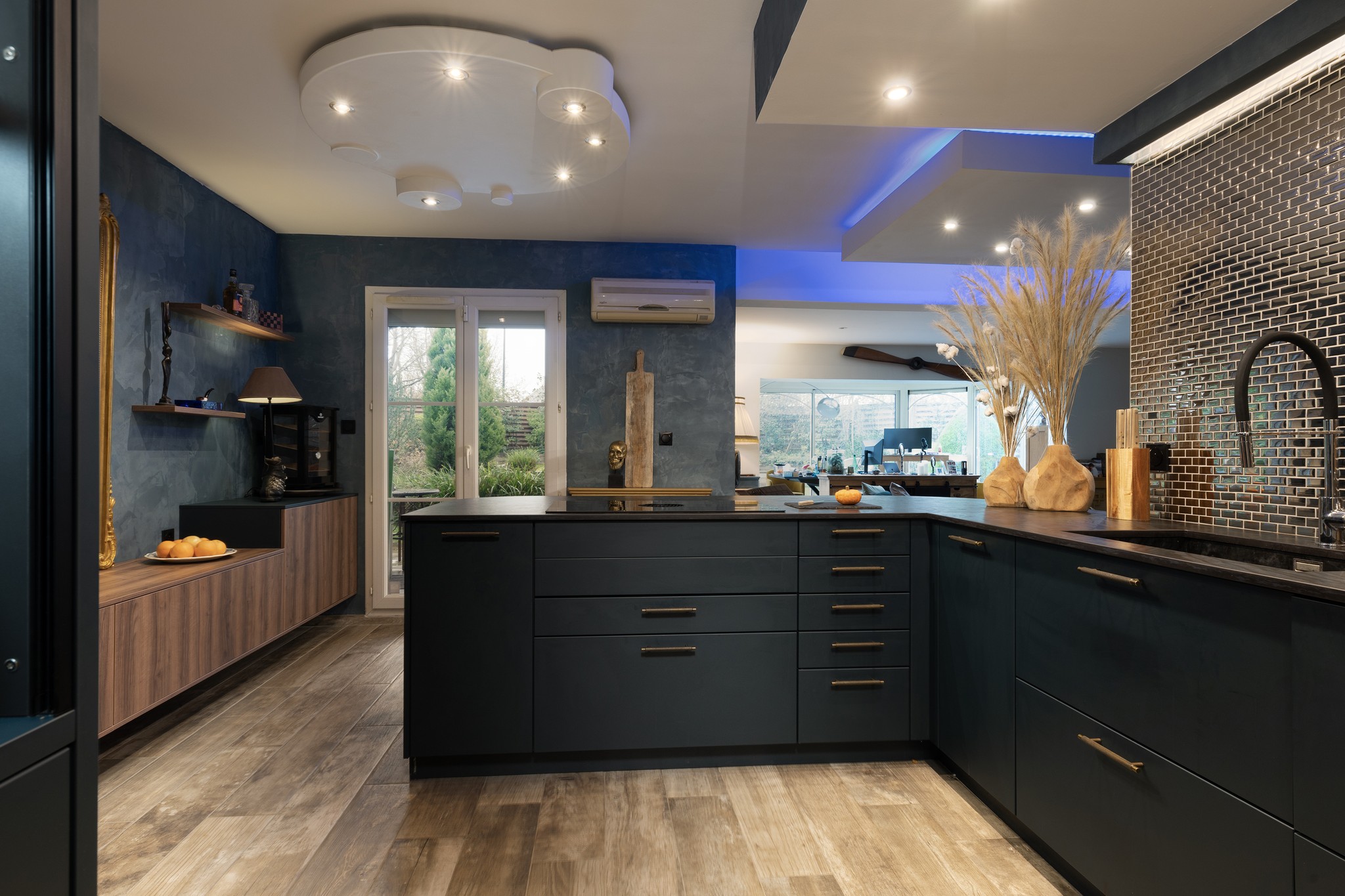 Cuisine moderne avec placards sombres, sol en bois, dosseret en mosaïque et grand îlot. La lumière naturelle pénètre par les portes vitrées. Des lumières décoratives au plafond et des éclairages d'appoint bleus ajoutent de la chaleur à l'espace.