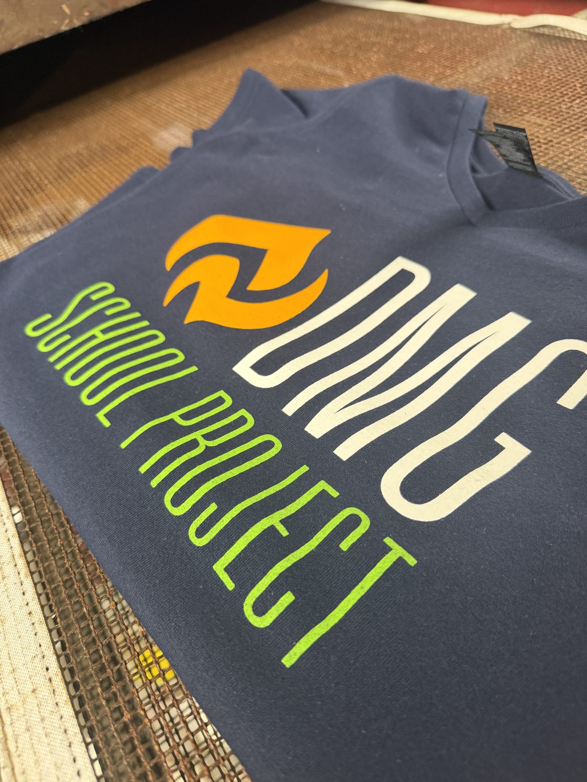 A blue-gray shirt with white and green DMG School Project lettering and an orange symbol.