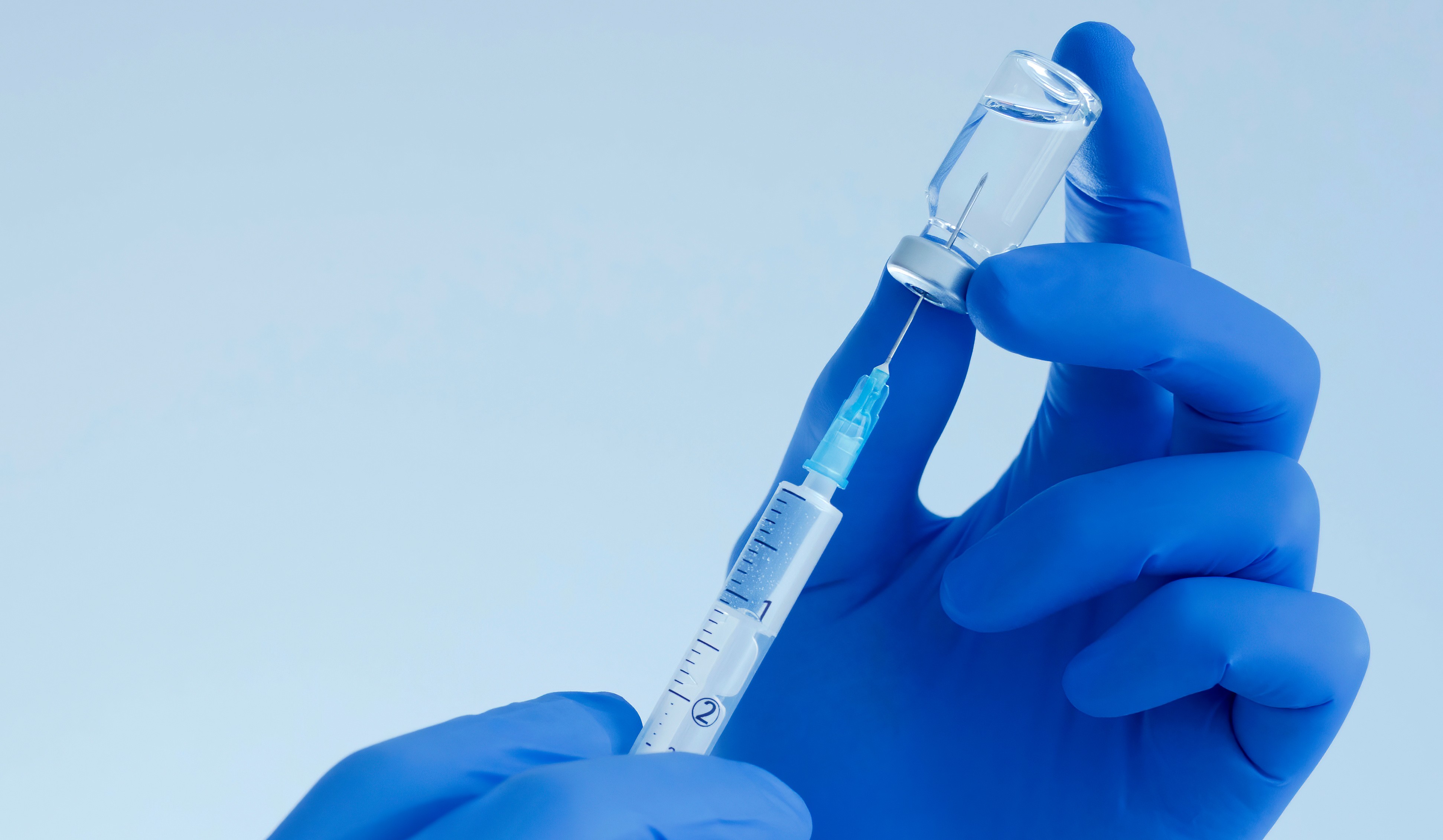 Hand wearing blue gloves, extracting a liquid out of a vial using a syringe.