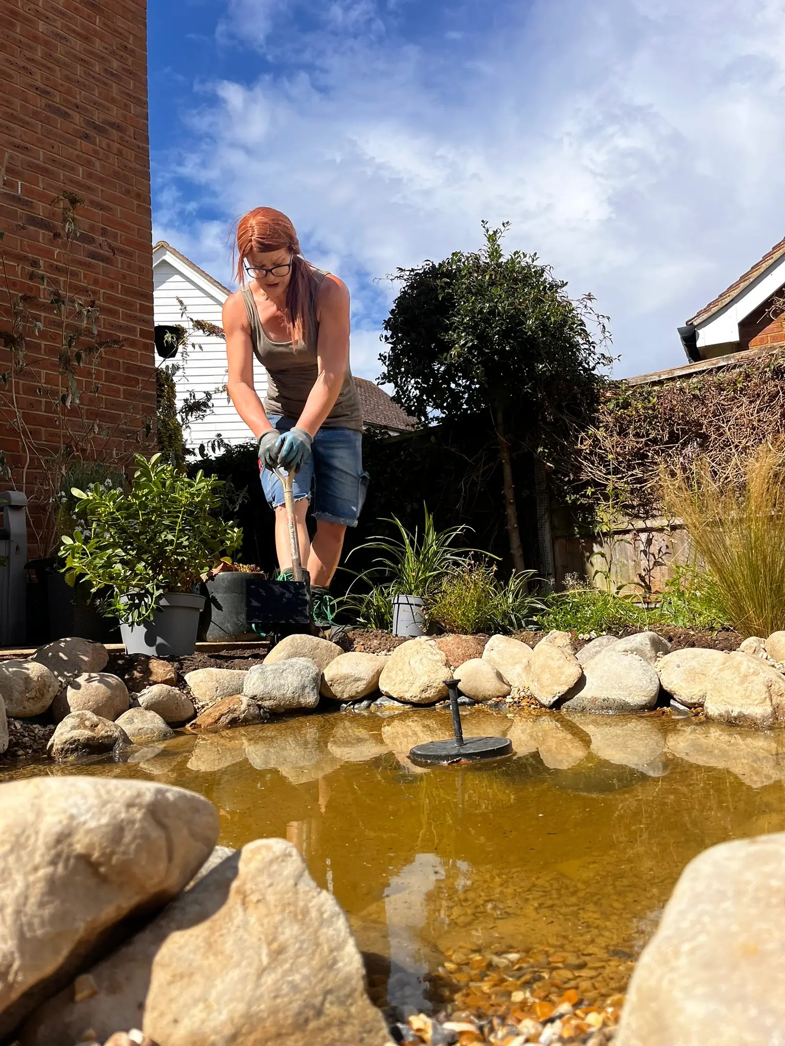 A person jumps over a pond surrounded by rocks and greenery, with a house and blue sky in the background.