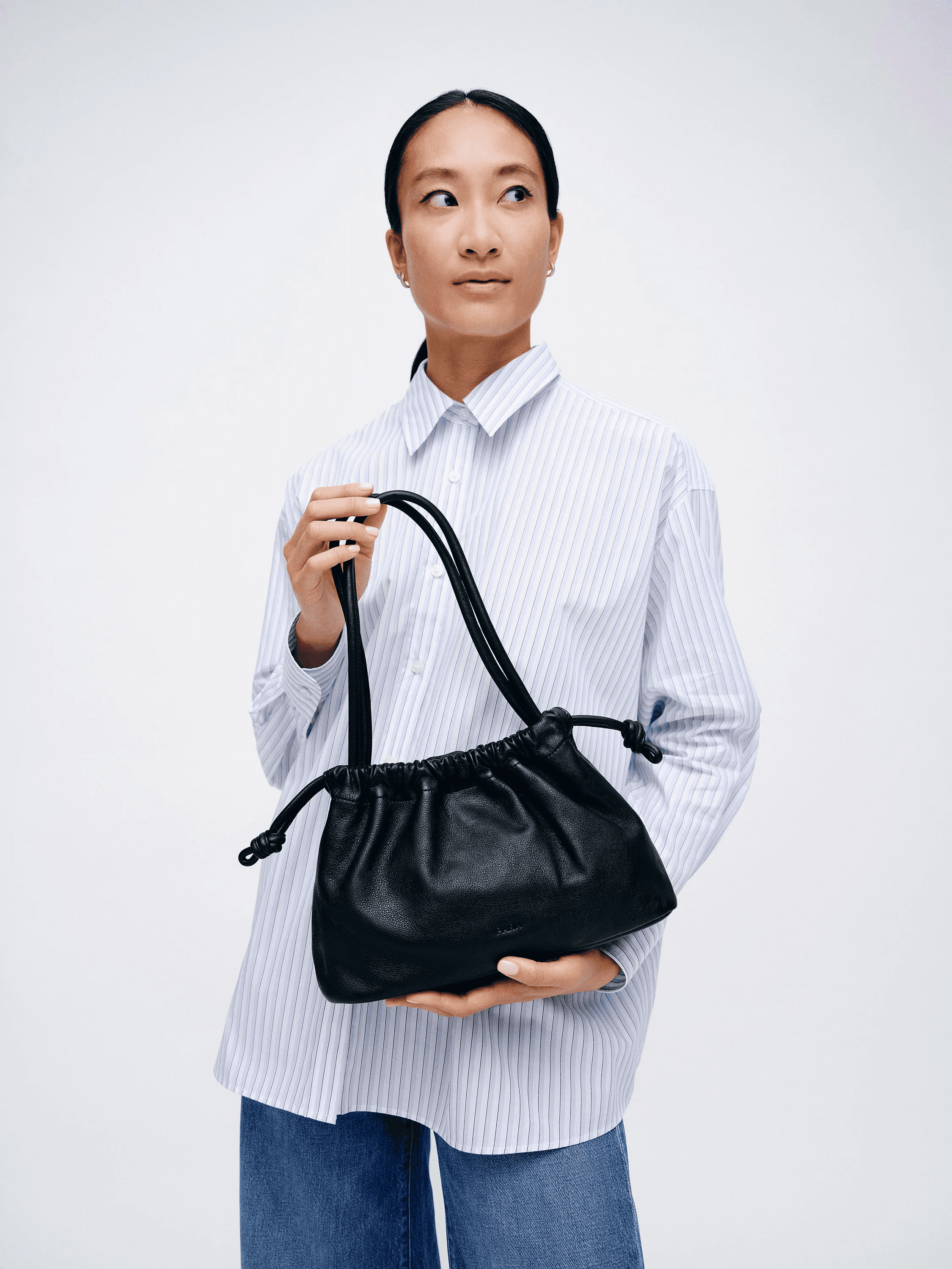 A person wearing a blue striped shirt and jeans holds a black leather handbag with drawstring details.