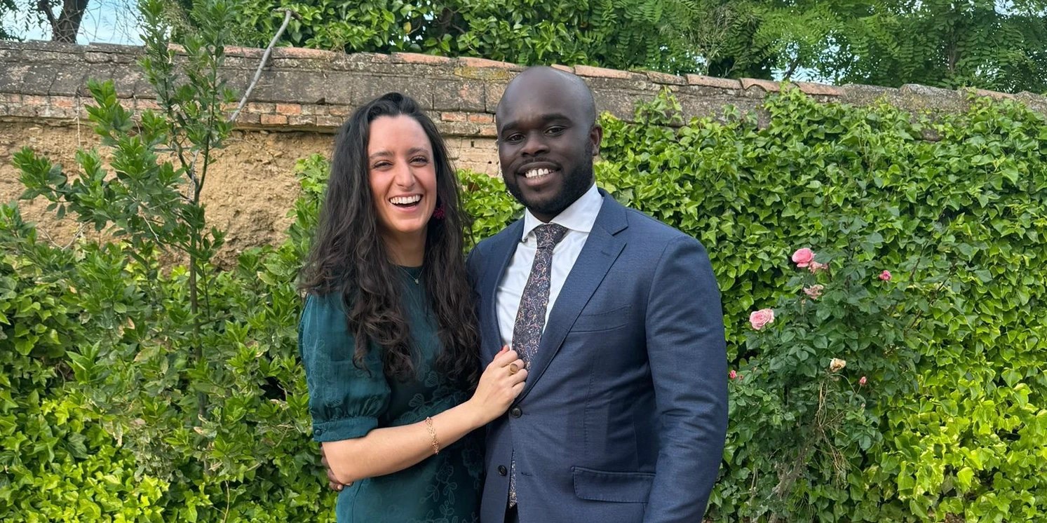 A bride and groom hold hands tenderly, the bride's bouquet of dried flowers hanging low. They stand in a softly lit, elegant interior, conveying romance.
