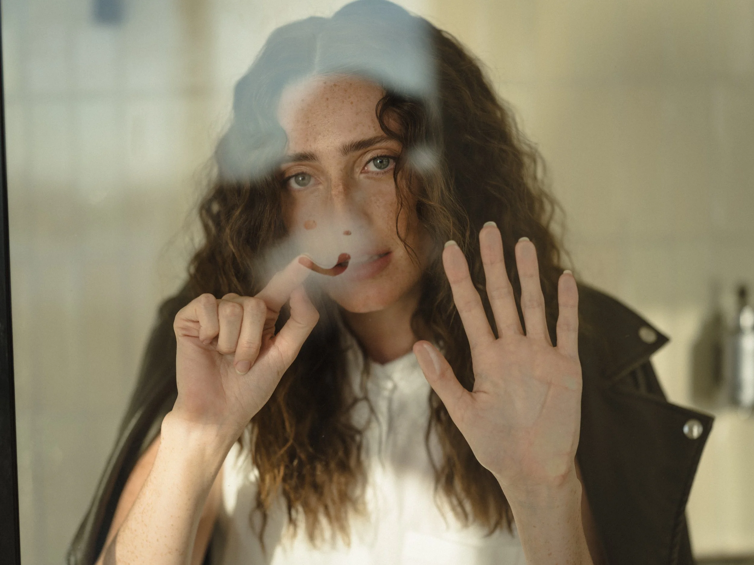 Woman with curly hair pressing her hand to a pane of glass, freckles catching warm light, leather jacket on her shoulders.