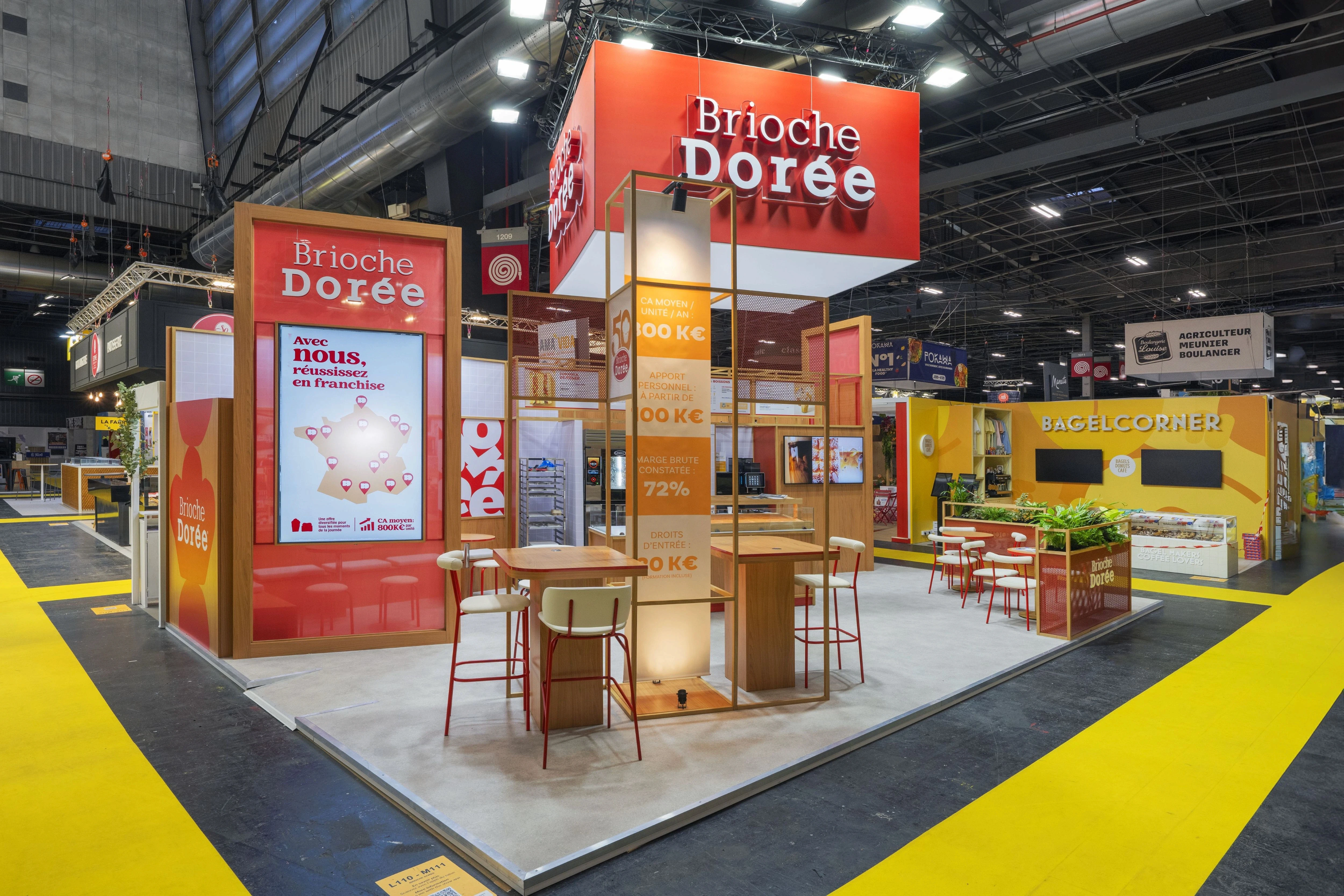 Stand Brioche Dorée conçu et réalisé par Expace pour Franchise Expo Paris 2026 – stand sur-mesure avec enseigne suspendue rouge, espace dégustation, comptoir boulangerie et tables d'accueil franchise.
