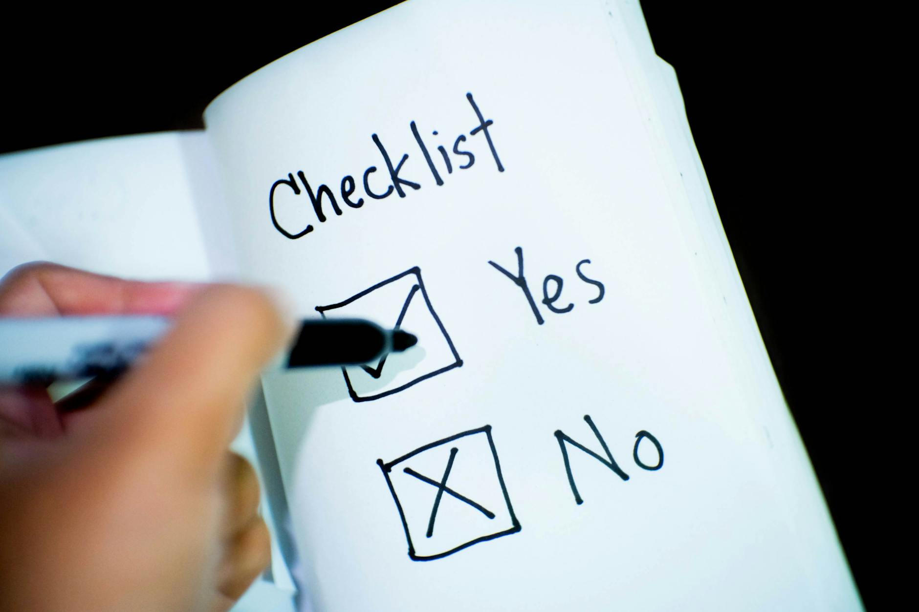 A hand checking off boxes on a printed checklist with a highlighter and a pair of glasses nearby.
