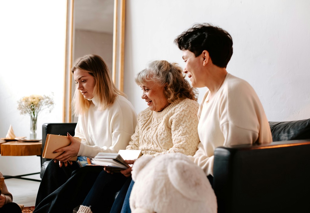 Three generations of women, dressed in cozy sweaters, enjoy reading and spending quality time together in a bright, comfortable living room.