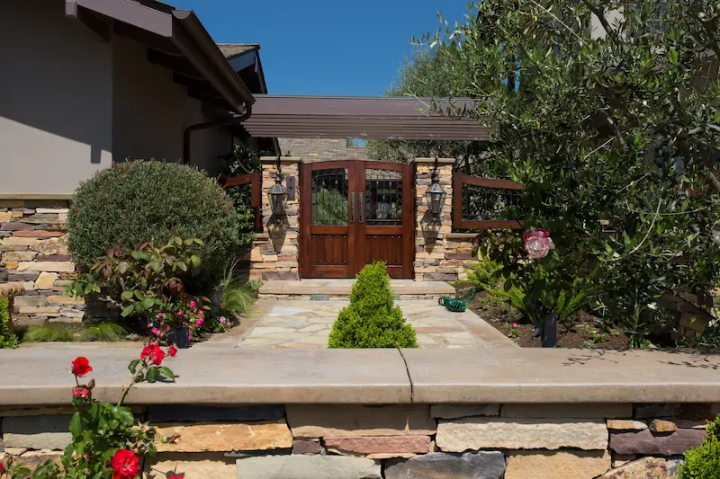 Lush patio entrance with dark wooden gate doors and stone hardscape. From the Big Canyon Remodel project. Photo by Chris Darnall.