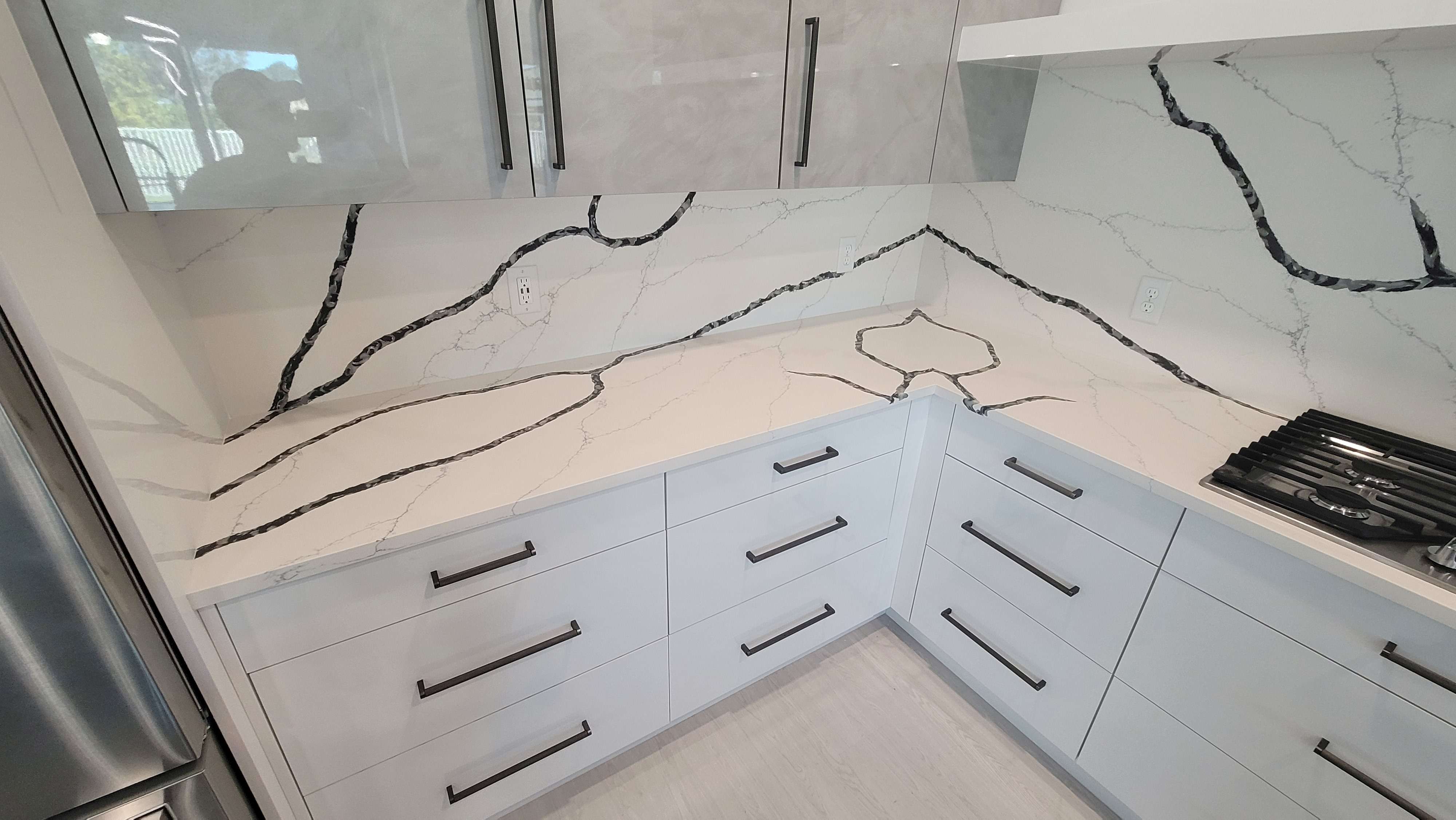 Corner of a kitchen showing high gloss white base cabinets, with ash gray handles, calacatta laza night countertop and backsplash, and high gloss gray wall cabinets