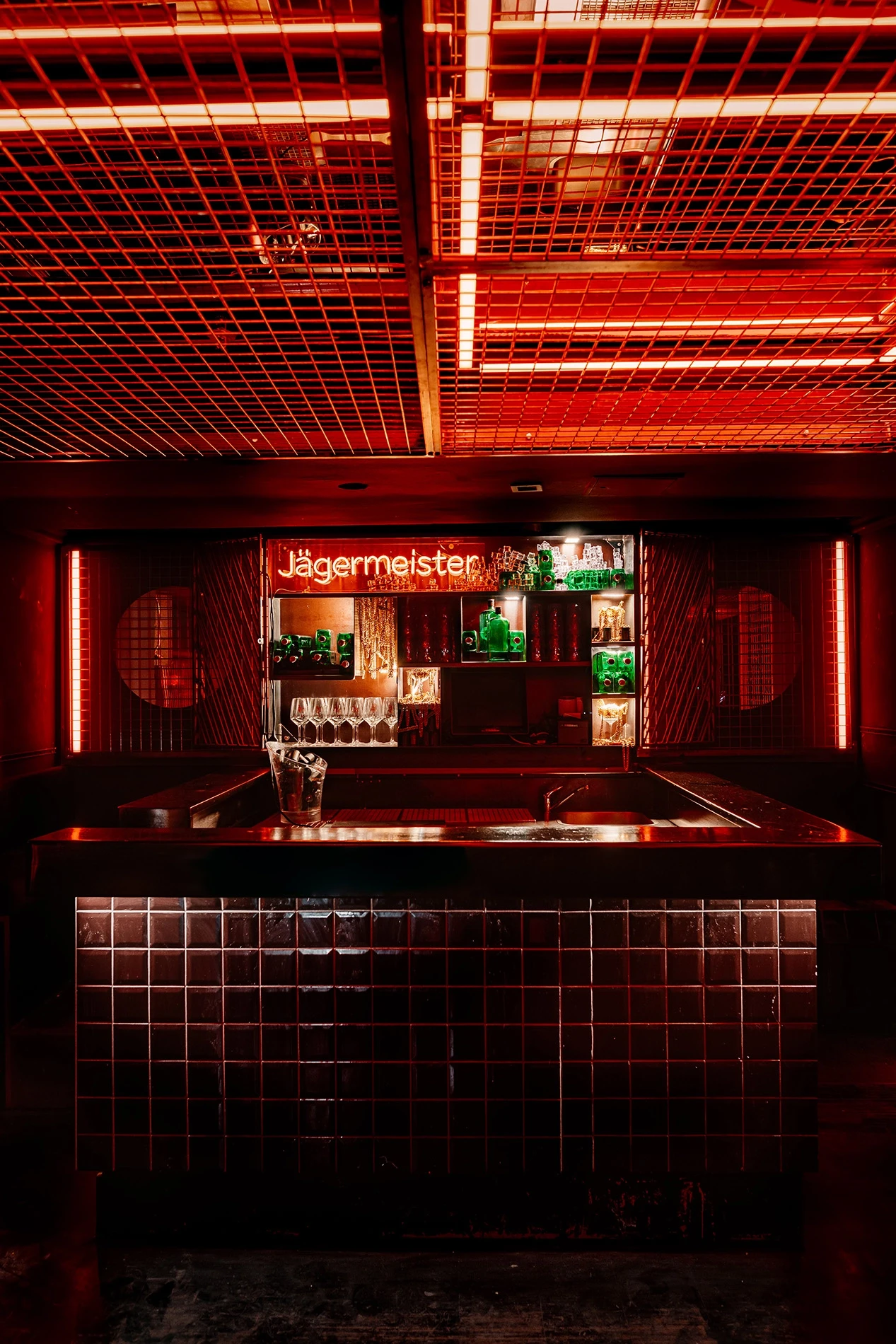 Red-lit bar at JÄGERMEISTER x CALL ME DRELLA with dark tiled front, an orange Jägermeister neon sign above backlit green bottles and a glowing grid ceiling.