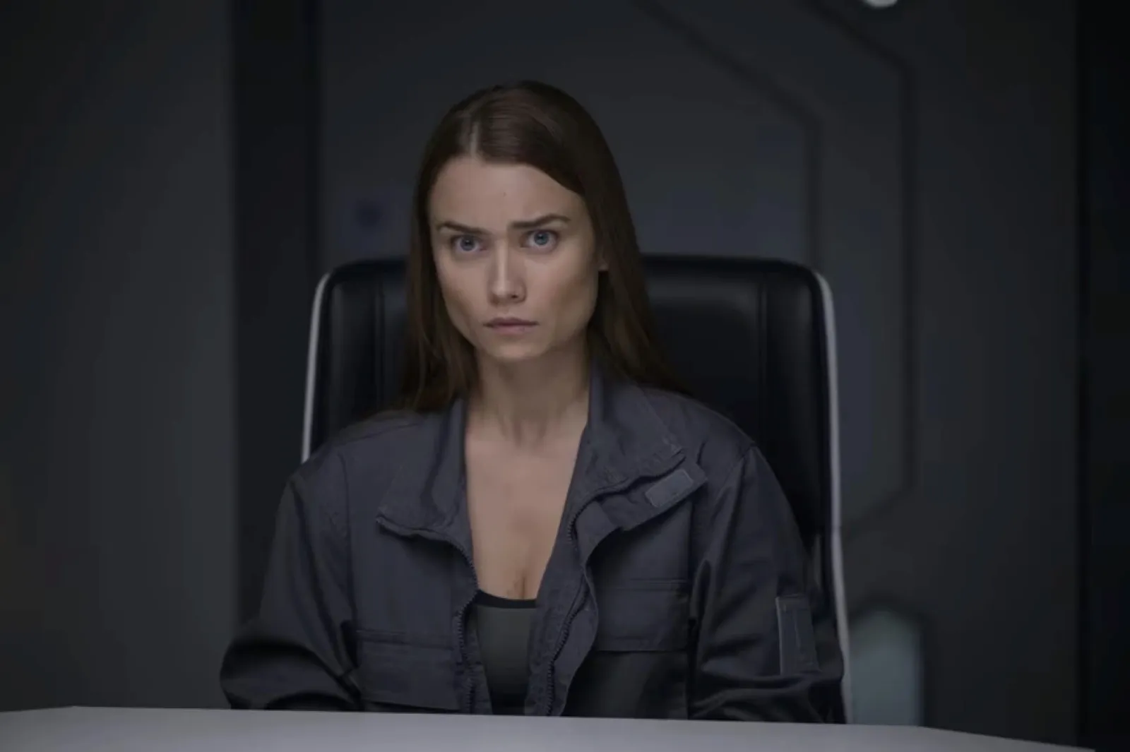 A woman with long brown hair sits at a table in a dimly lit futuristic room, wearing a dark jacket, and looking intently forward, conveying a serious or intense expression.