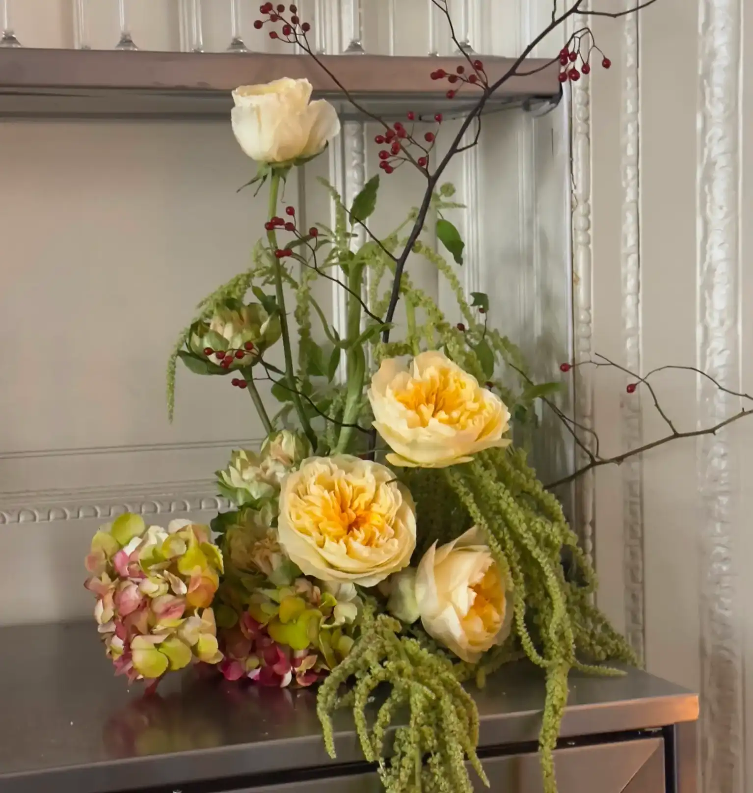 Arreglo floral de estilo contemporáneo con rosas color crema y centros amarillos, hortensias verdes y rosadas, ramas de bayas rojas y tallos colgantes de amarantos, dispuesto sobre una superficie metálica frente a una pared blanca con molduras decorativas.