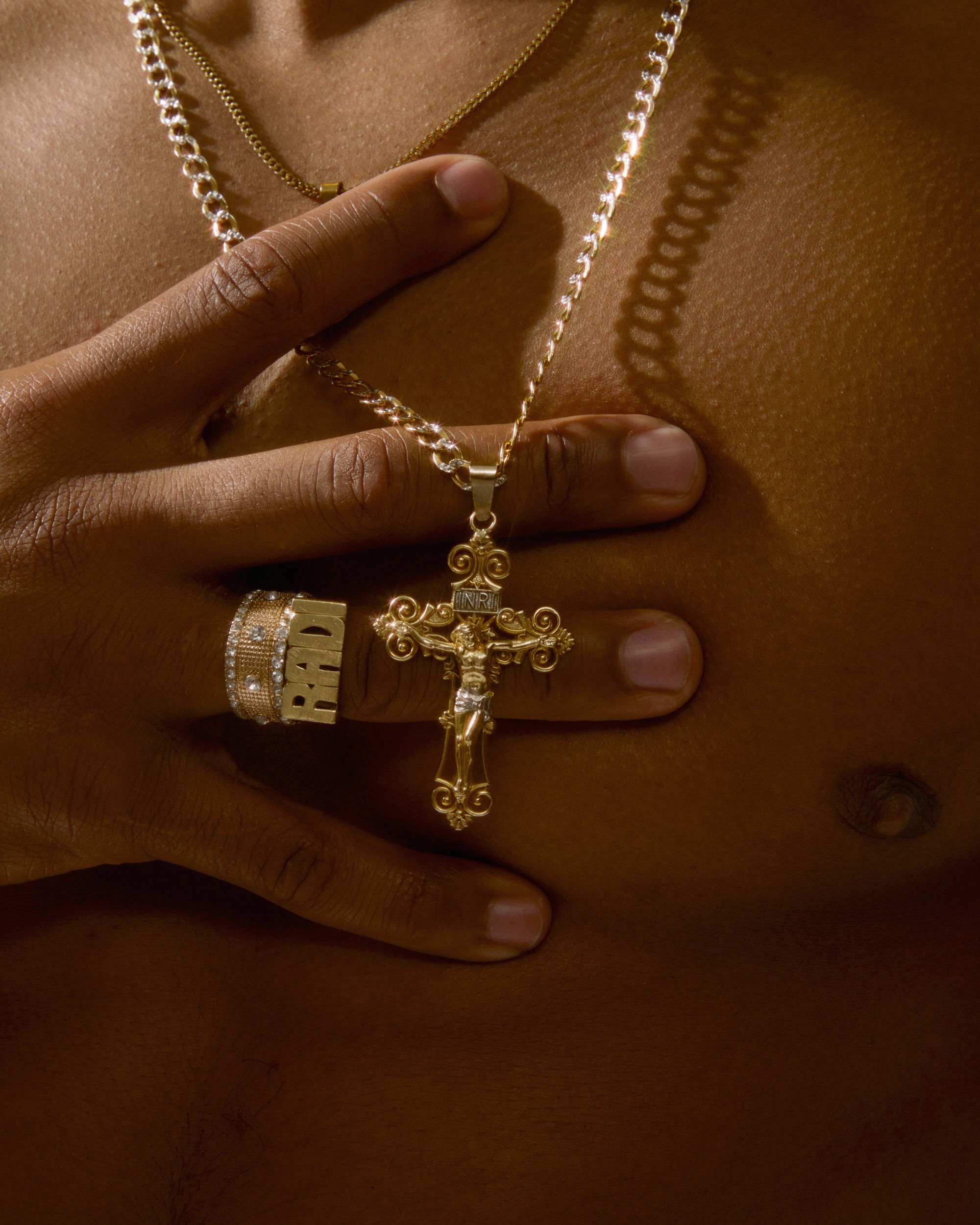 A close-up of a man's hand holding a gold crucifix pendant against bare skin, a diamond-encrusted ring visible on his finger.