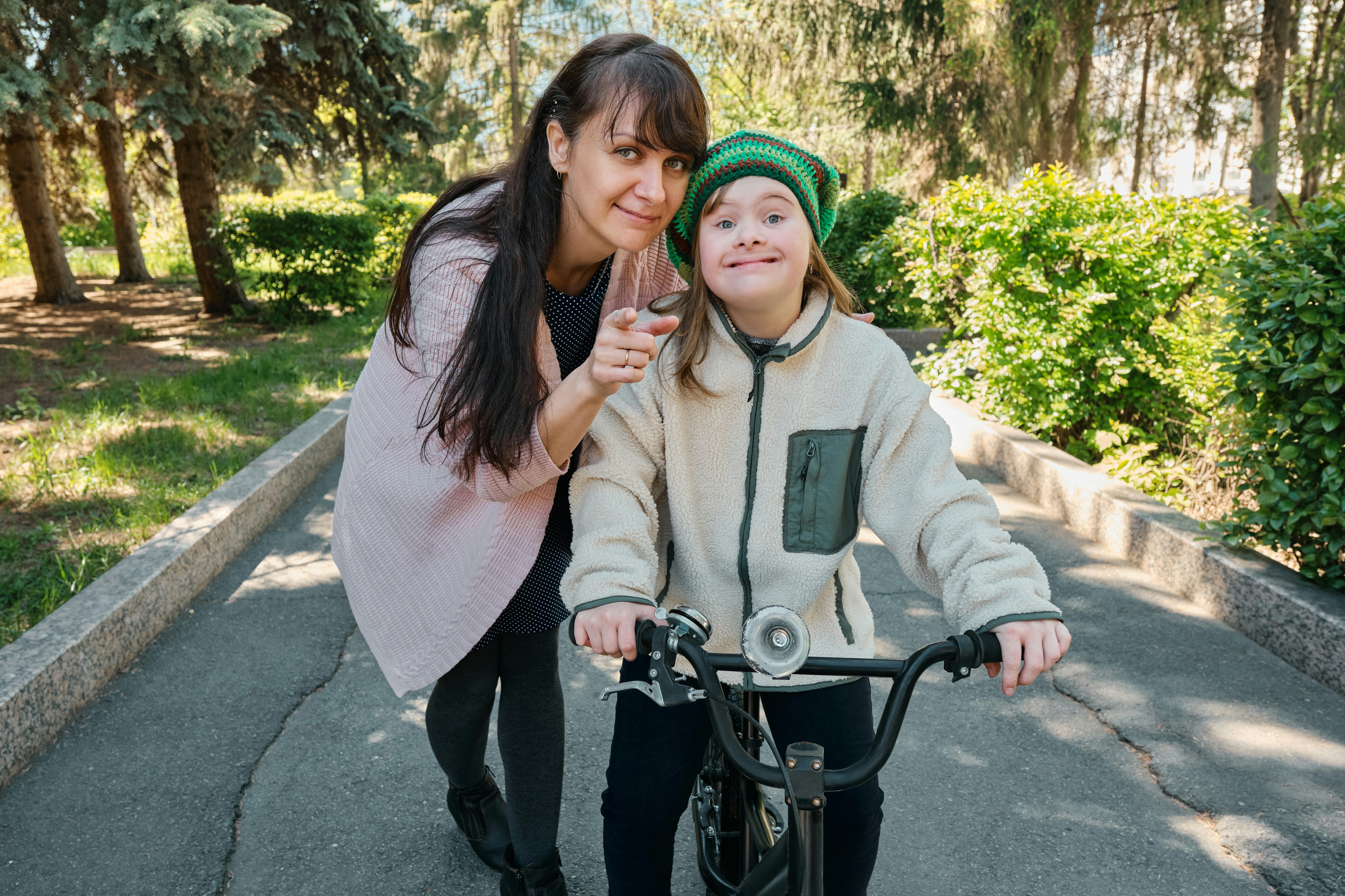 Woman and child on a bike path in a park. The woman stands beside a child on a bike, smiling and pointing. The child wears a green helmet and beige jacket.