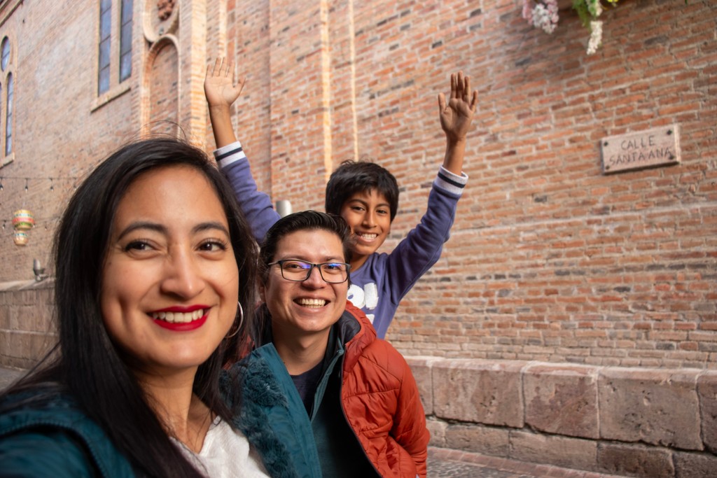 A group of three people smiling and posing in front of a rustic brick wall with a street name sign reading "Calle Sangratana," capturing a joyful and relaxed moment in an urban setting.