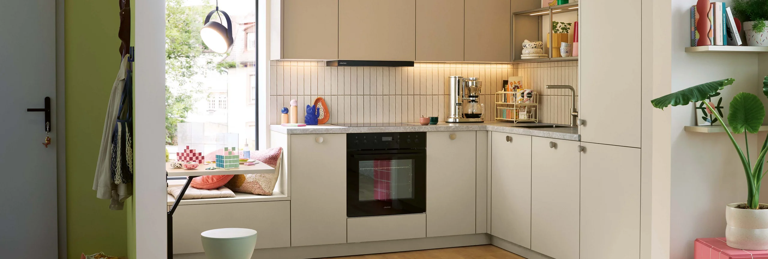 Cuisine en L Schüller SORA aux façades beige amande, avec rangements hauts et coin repas près de la fenêtre