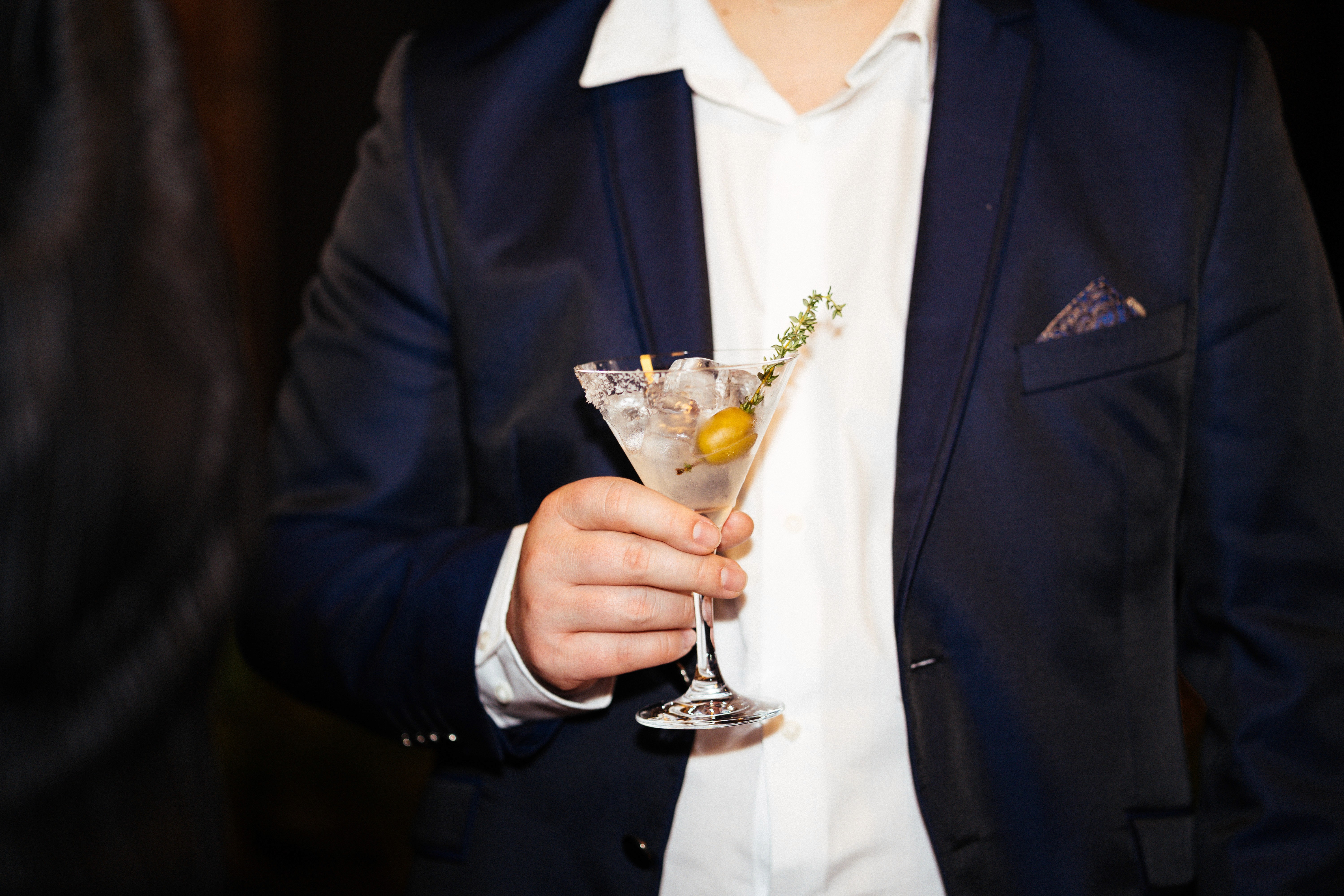 A person in a dark suit holds a cocktail glass, with a white shirt visible underneath the jacket.