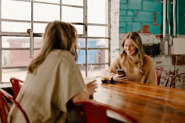 Two women sitting at a wooden table in a cafe, one using her phone while talking.