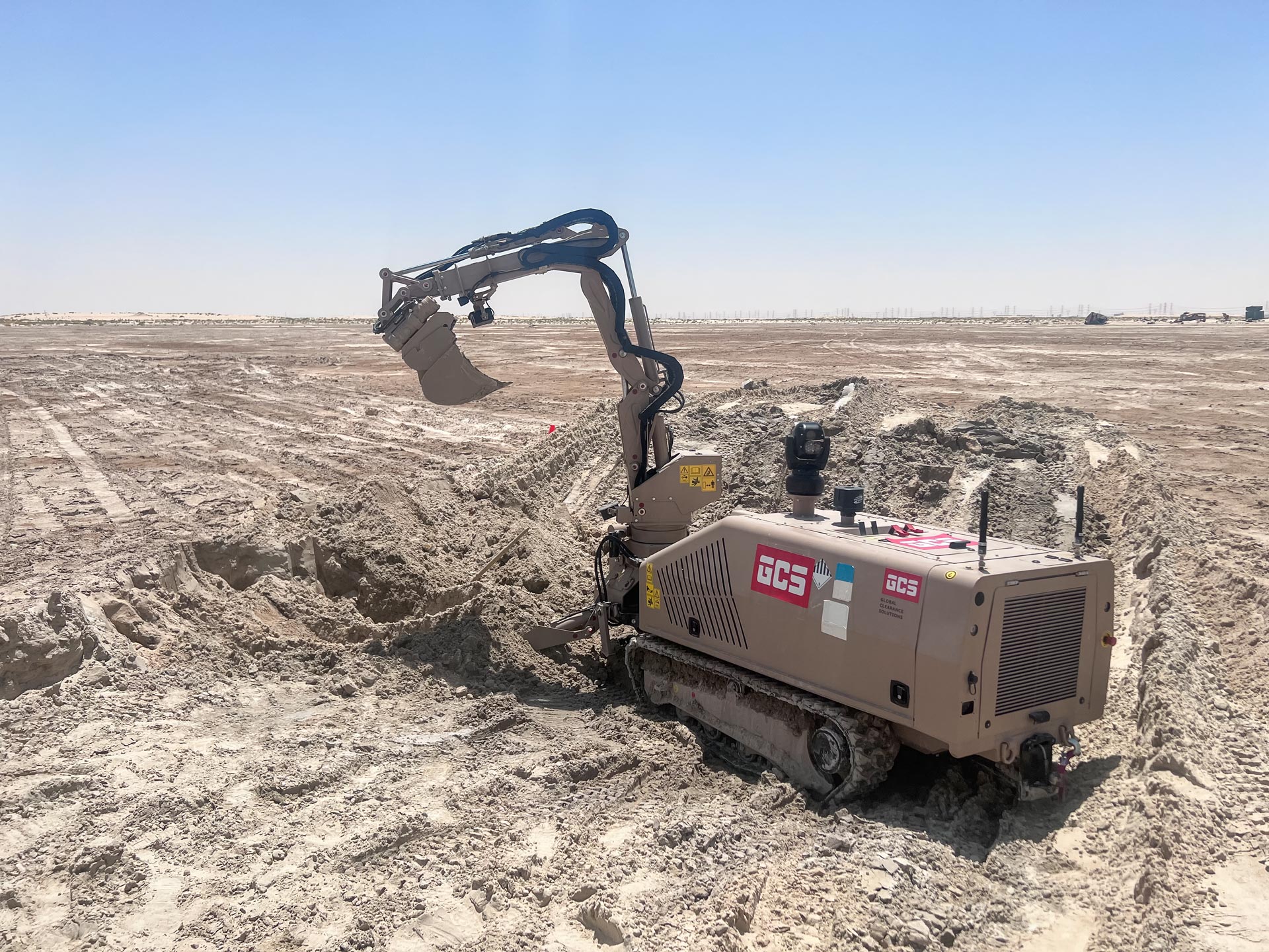 GCS-100 remote-controlled platform with manipulator arm and bucket tool excavating during range clearance operations in a desert environment.