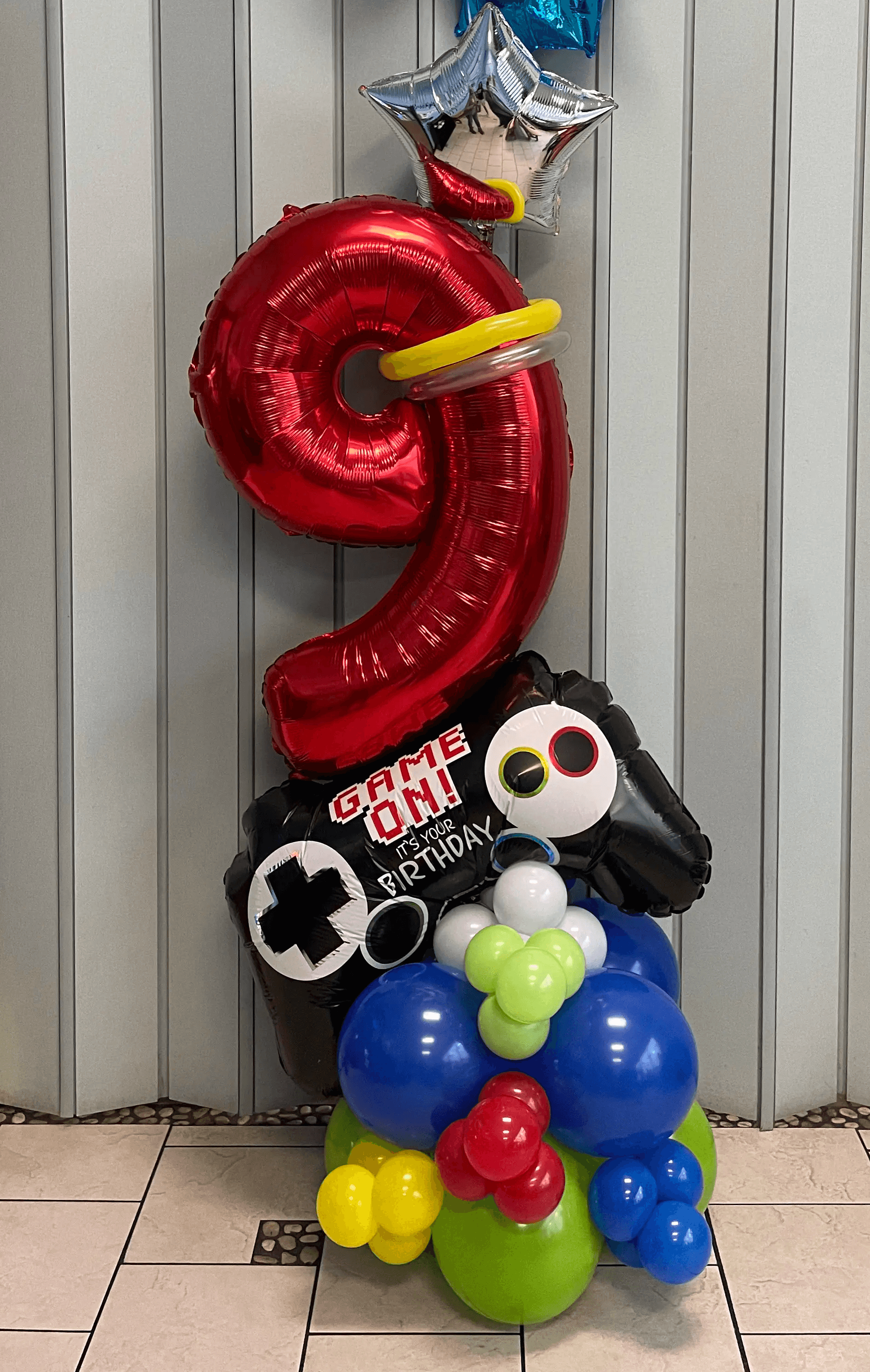 Gamer-themed birthday balloon display featuring a large red number “9” balloon, a “Game On” controller design, and bright, playful balloon clusters in blue, green, yellow, and red. Perfect for young gaming enthusiasts. Susa’s Sweets & Balloons.