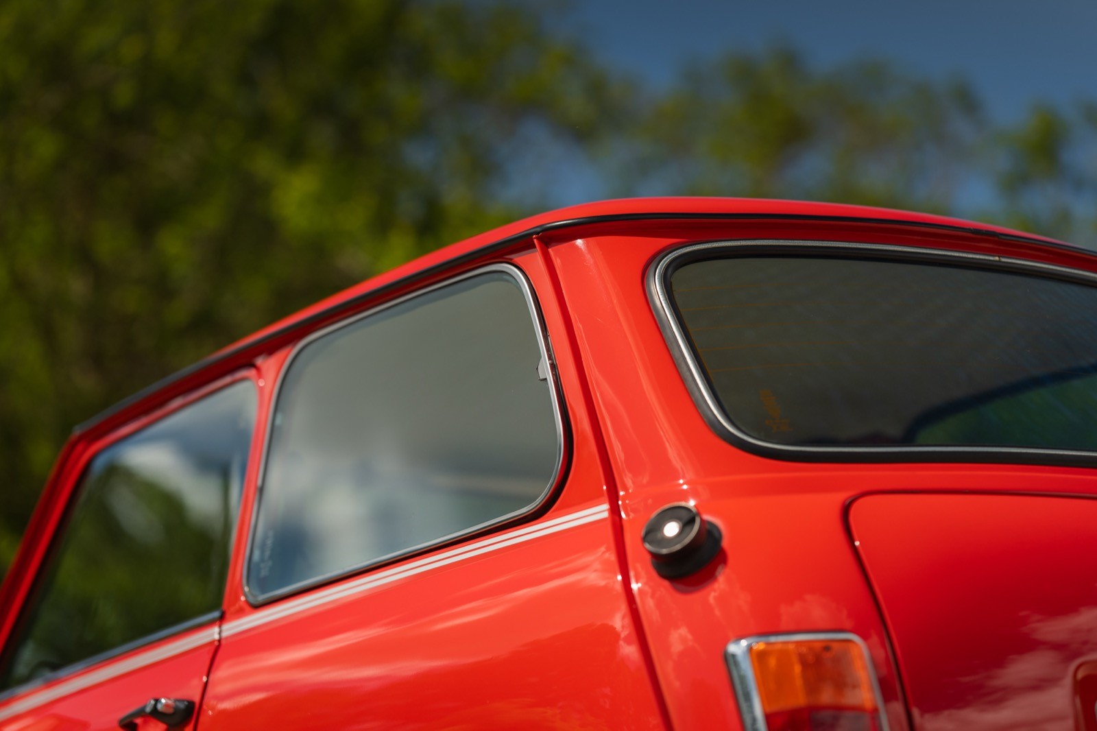 A close-up of a red car, focusing on the rear window and part of the body, with greenery in the background.