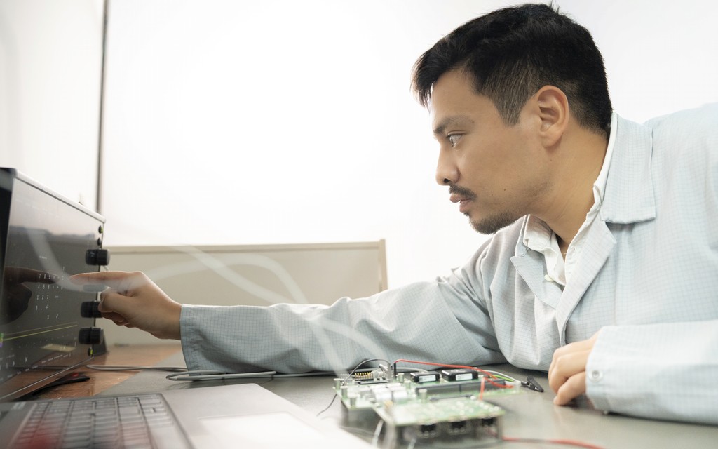 A man in a lab coat sitting at a desk, with a circuit board and wiring in front of him, a laptop, and adjusting a device with a knob on the desk with his index finger.