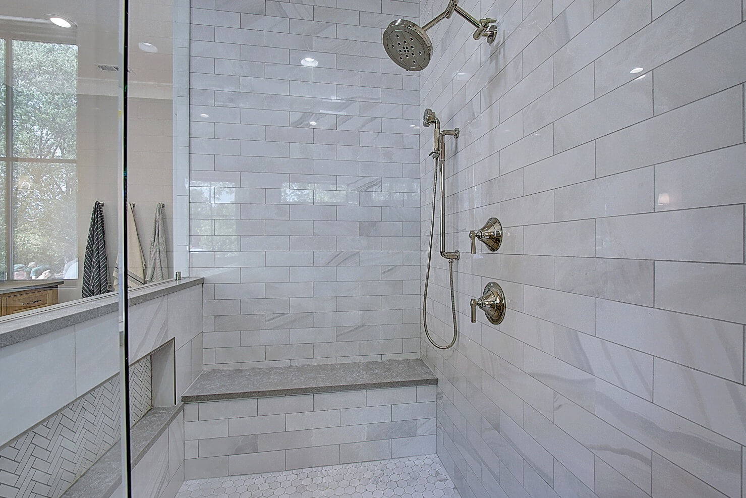 Modern bathroom shower with sleek marble tiles, two showerheads, and a large glass door. Natural light streams in.
