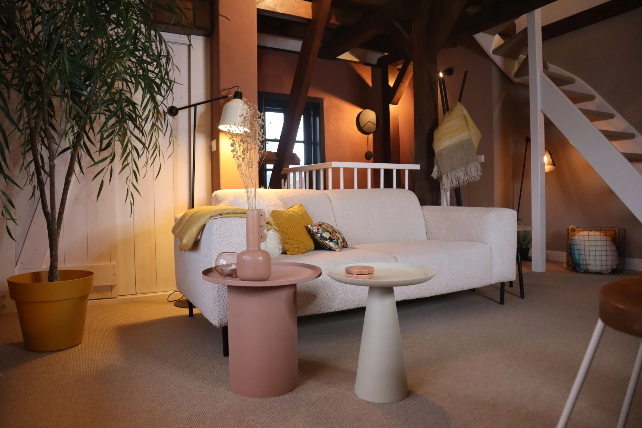 Gezellige woonkamer met een witte bank, twee pastelkleurige bijzettafels, een grote kamerplant in een gele pot en zachte verlichting. De ruimte heeft een minimalistisch ontwerp met houten balken en een open trap.