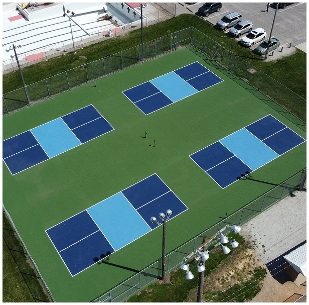 Aerial view of four blue pickleball courts on a green surface surrounded by fencing at a sports facility