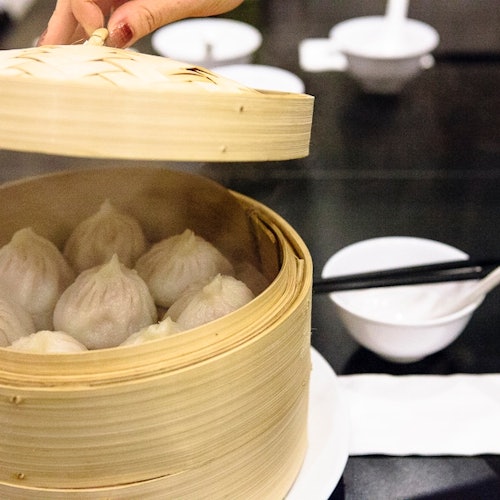 一只手将竹蒸笼的盖子掀开，露出了蒸好的饺子。白色桌布下放着筷子和碟子。