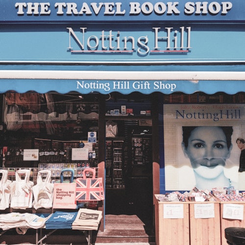 Storefront of "The Travel Book Shop" and "Notting Hill Gift Shop" with bags, books, and a large "Notting Hill" movie poster.