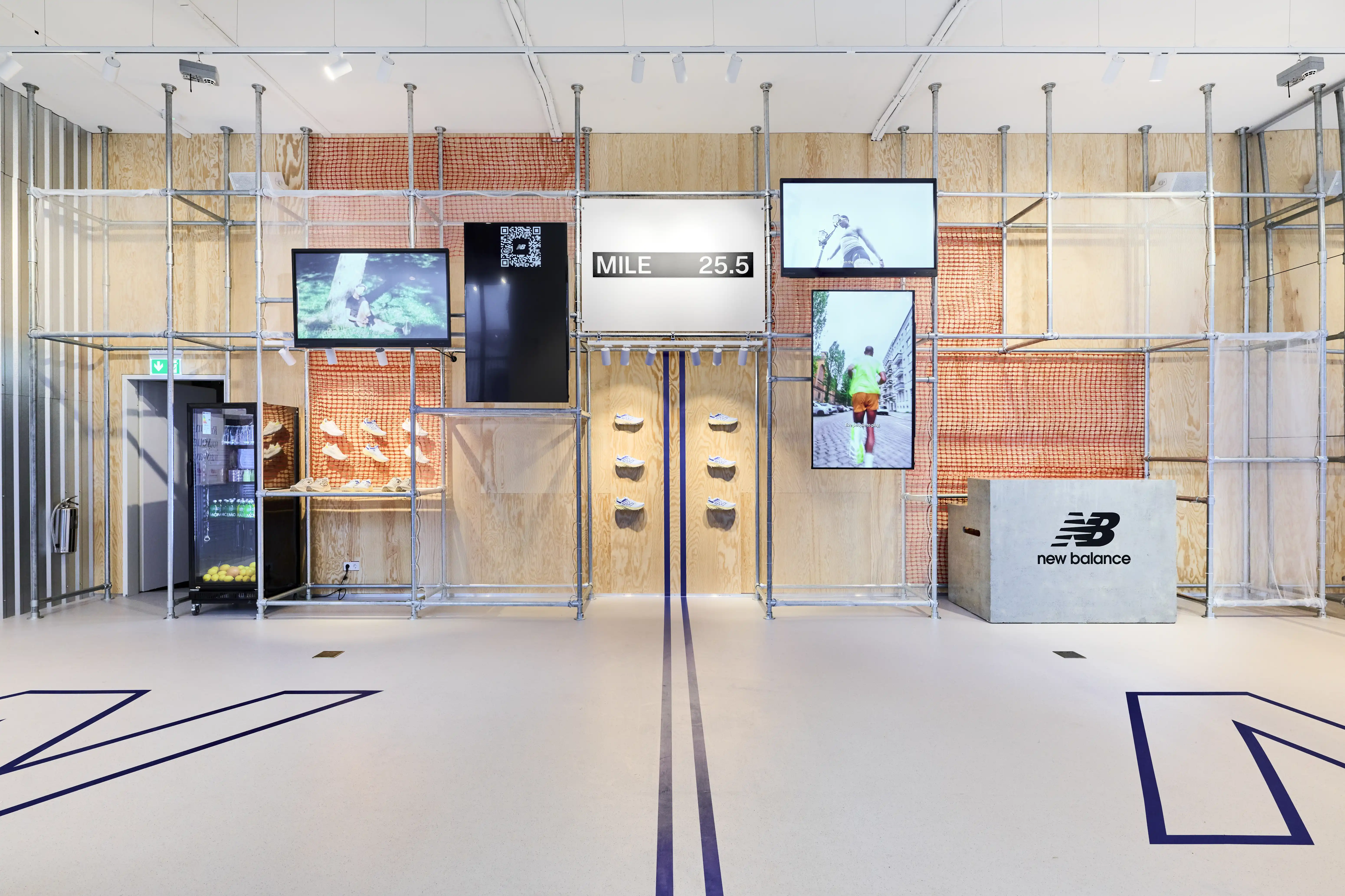 Scaffold wall with screens, orange safety netting and shoe displays at the Highsnobiety x New Balance pop-up, with a blue line running across the floor.