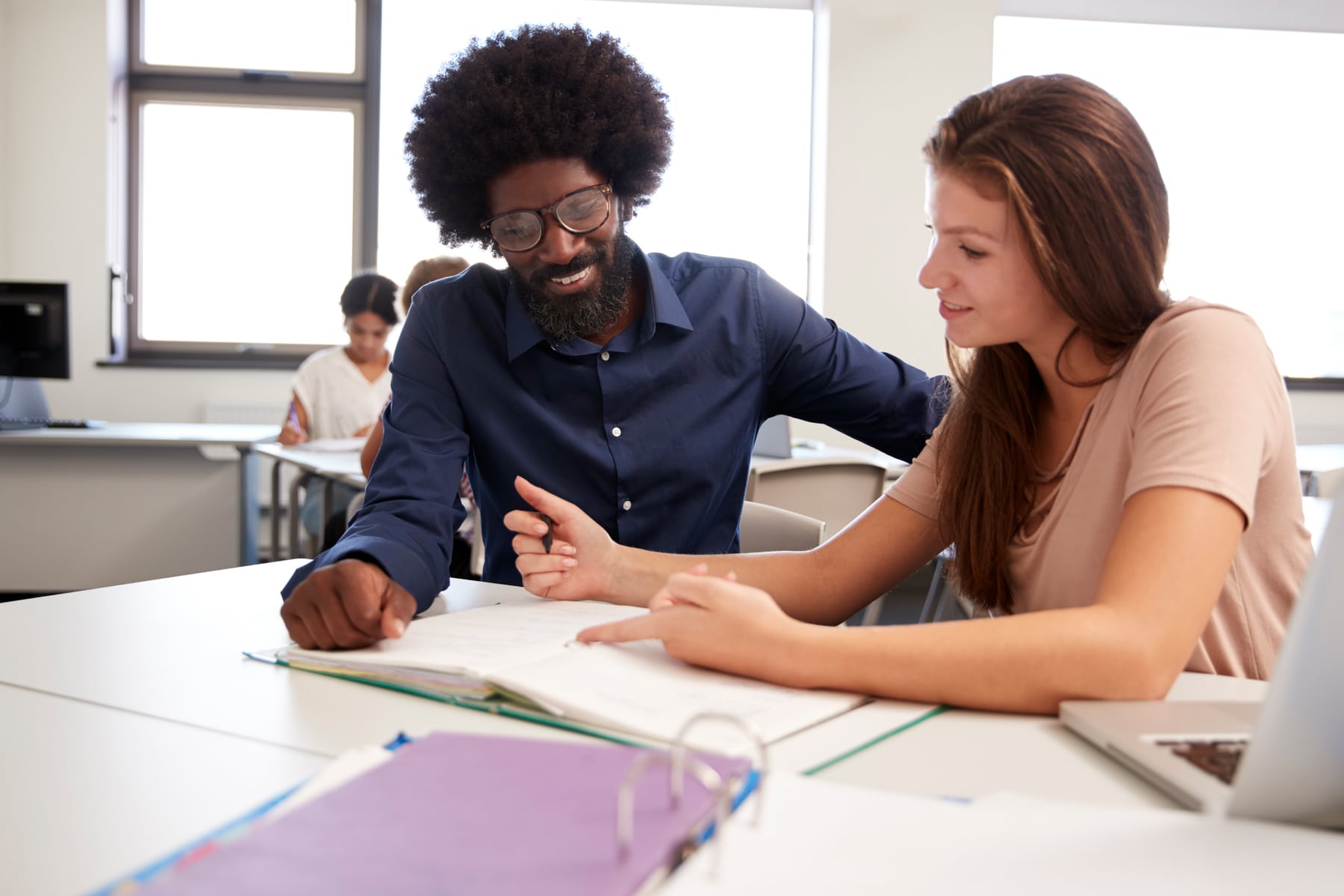 A teacher and student sit side by side in a classroom, looking over an open notebook as they discuss the work.