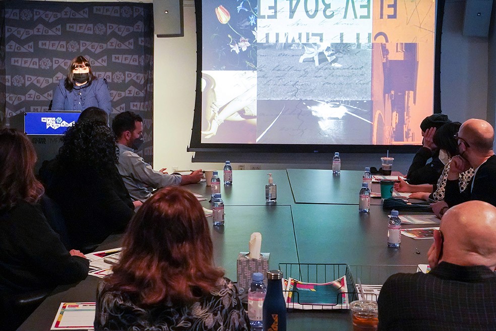 A woman dressed in a blue coat is giving a speech to the people sitting around a table, and on the right side of the wall is an image projected with a collage of a couple of images