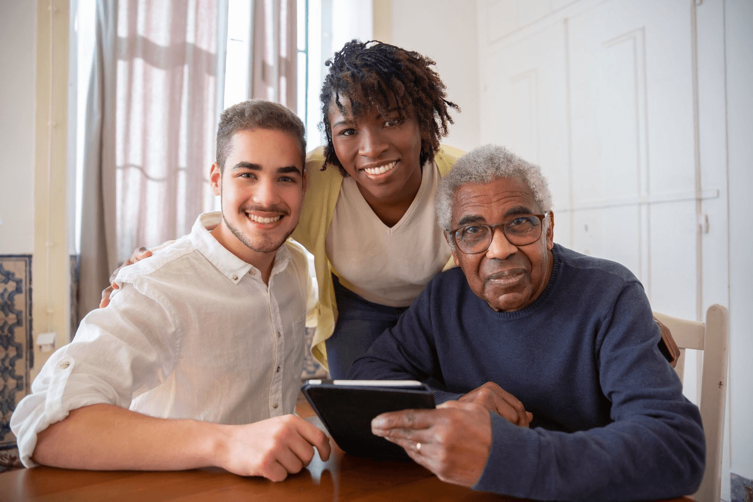 An older man shows a tablet to his daughter and grandson at the kitchen table.