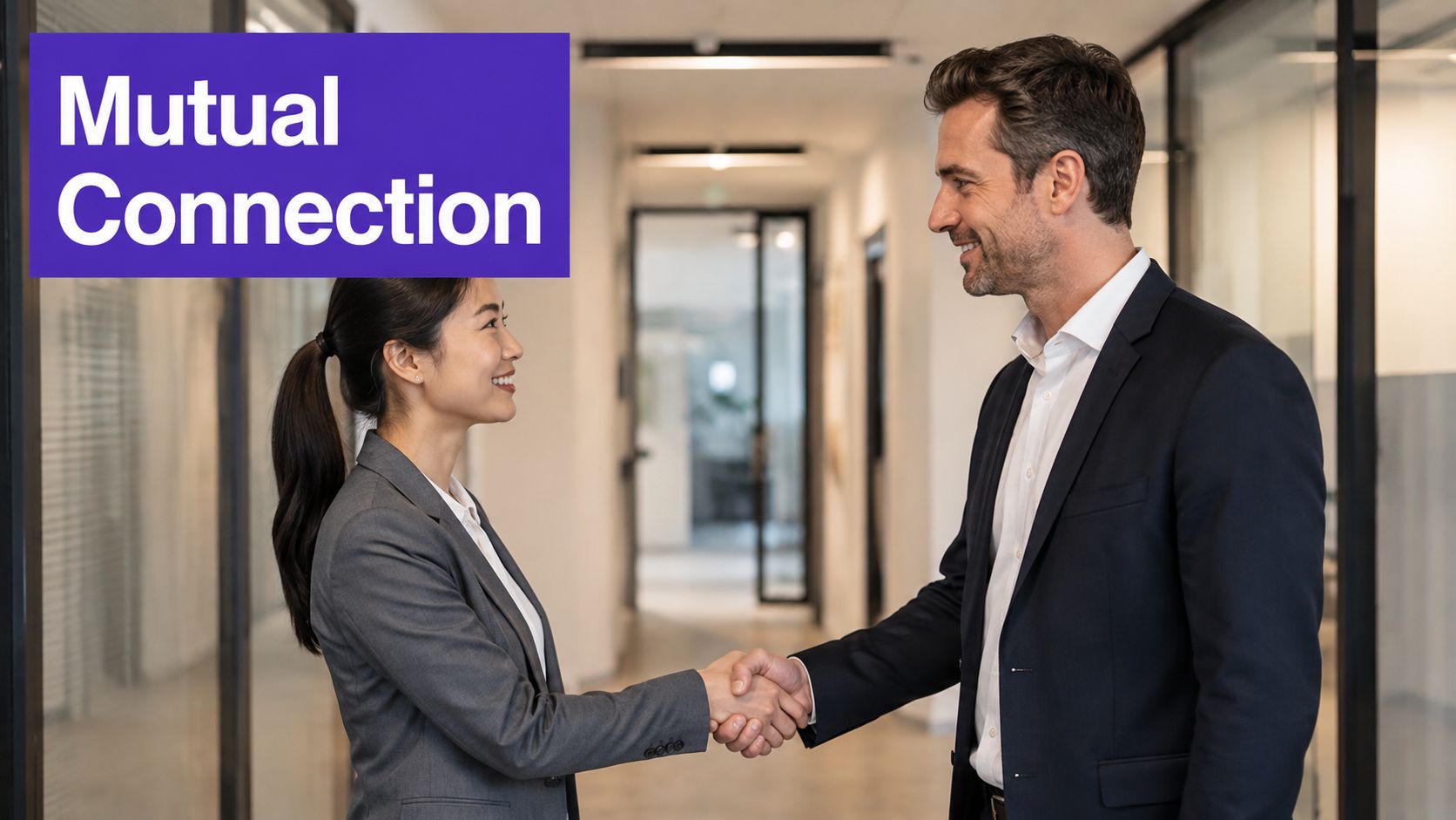 A professional man and woman in business attire shaking hands in a modern office hallway.